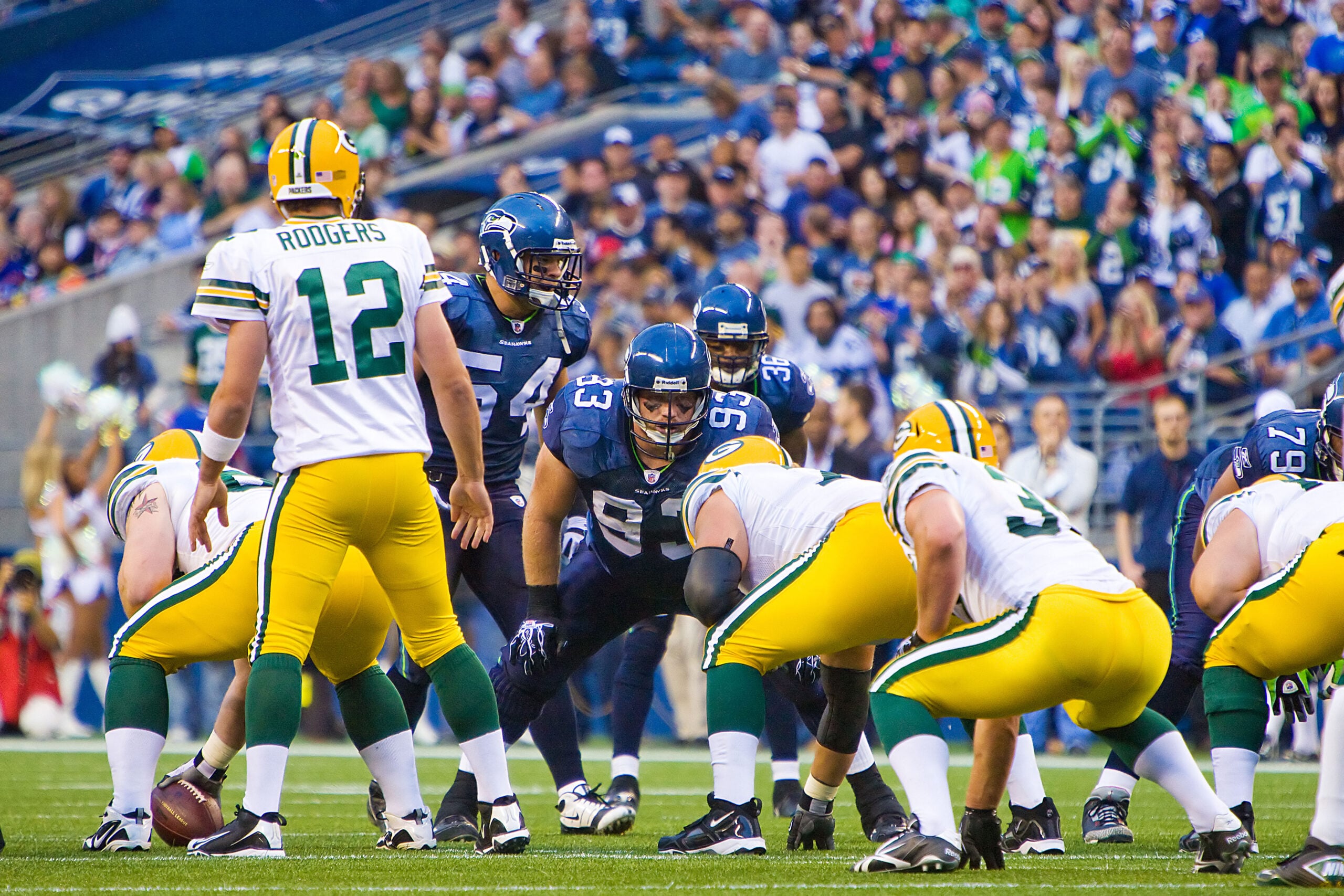 Os Green Bay Packers e os Seattle Seahawks em um intenso confronto durante um jogo de futebol americano da NFL. Os jogadores estão em plena ação, alguns correndo com a bola, enquanto outros se posicionam defensivamente. O estádio está cheio de espectadores entusiasmados, ansiosos para acompanhar cada lance da partida. Os Packers estão vestindo seus tradicionais uniformes com as cores verde e amarelo, enquanto os Seahawks estão com seus uniformes nas cores azul e branco.