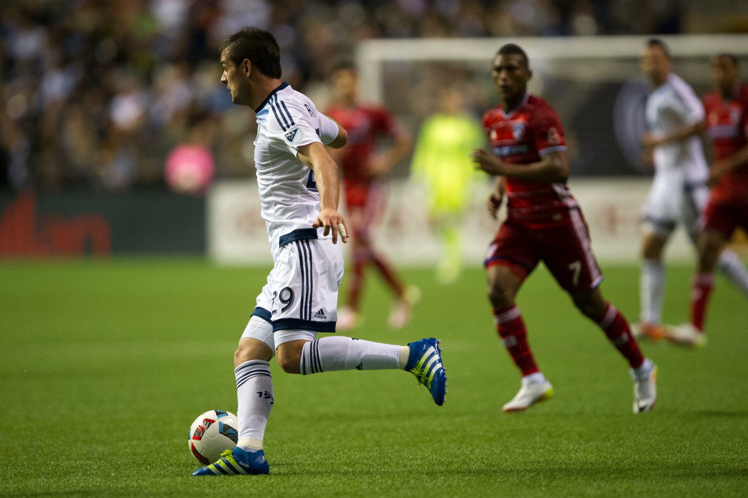A imagem mostra Octavio Rivero, atacante do time de Vancouver, correndo com a bola durante o jogo contra o Dallas pela MLS. Ele está vestindo uma camisa branca com listras azuis. Ele está dominando a bola. Ao fundo, é possível ver um campo de futebol e alguns espectadores nas arquibancadas.