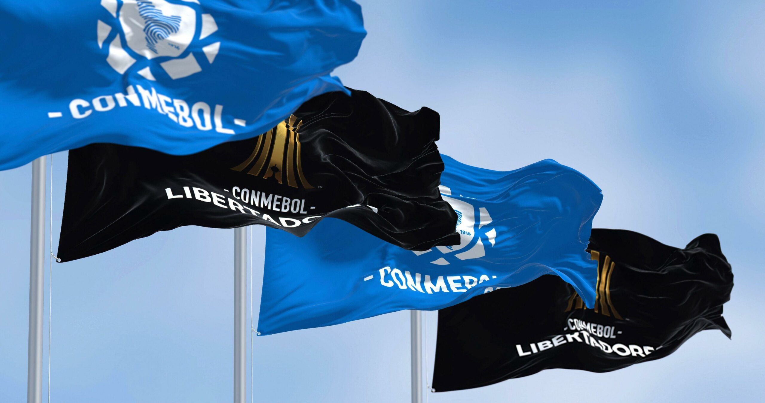 Imagem de uma fileira de bandeiras com os logotipos da CONMEBOL e da Libertadores tremulando ao vento. As bandeiras são nas cores azul e preto e estão dispostas em uma linha. Elas estão sendo içadas em um mastro, que é visível ao fundo.