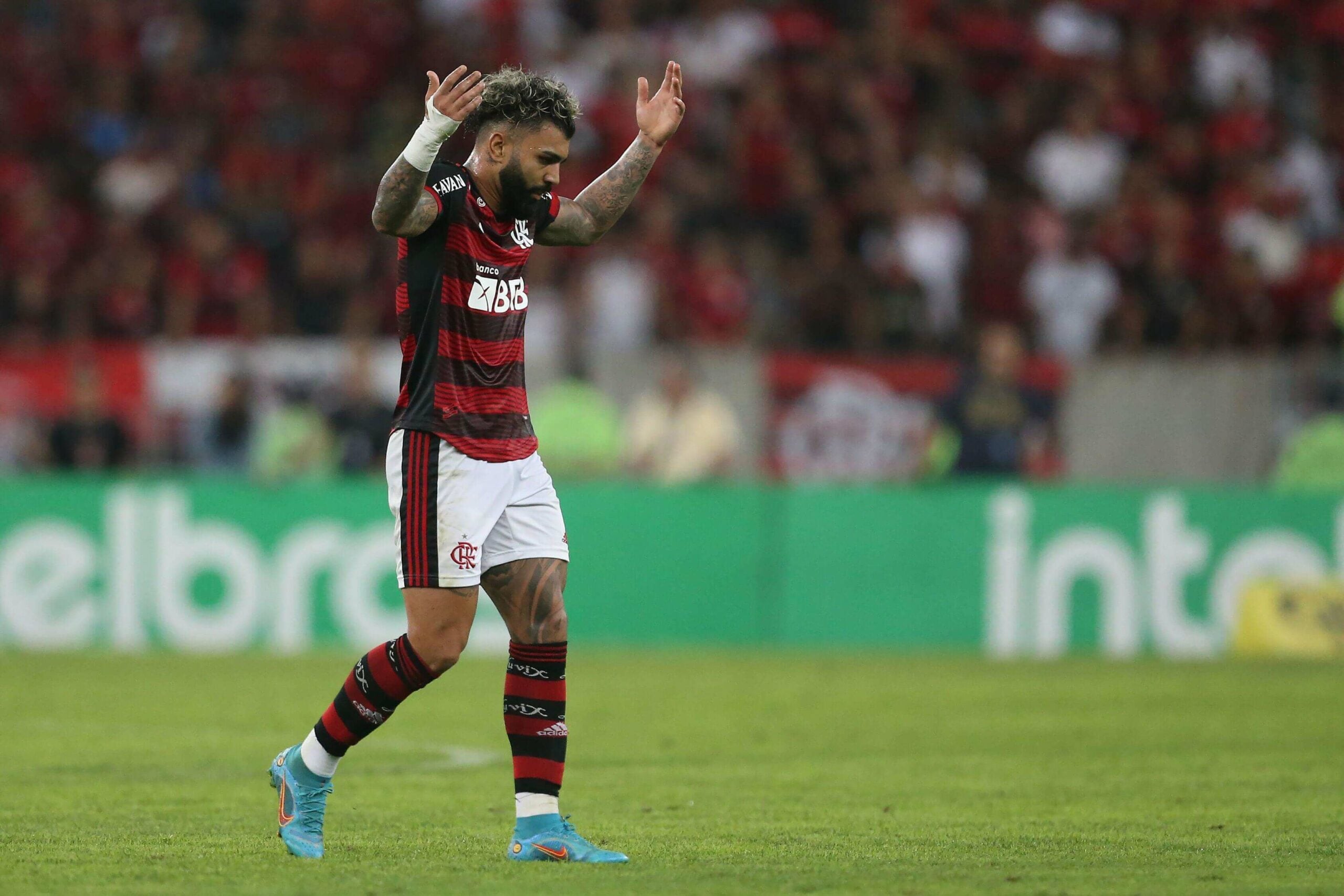 A imagem mostra Gabigol, jogador do Flamengo, com os braços levantados em comemoração após marcar um gol. Ele está vestindo a camisa do time e segurando uma bola de futebol. Ao redor do campo, é possível ver o estádio lotado com uma grande multidão de torcedores assistindo ao jogo.