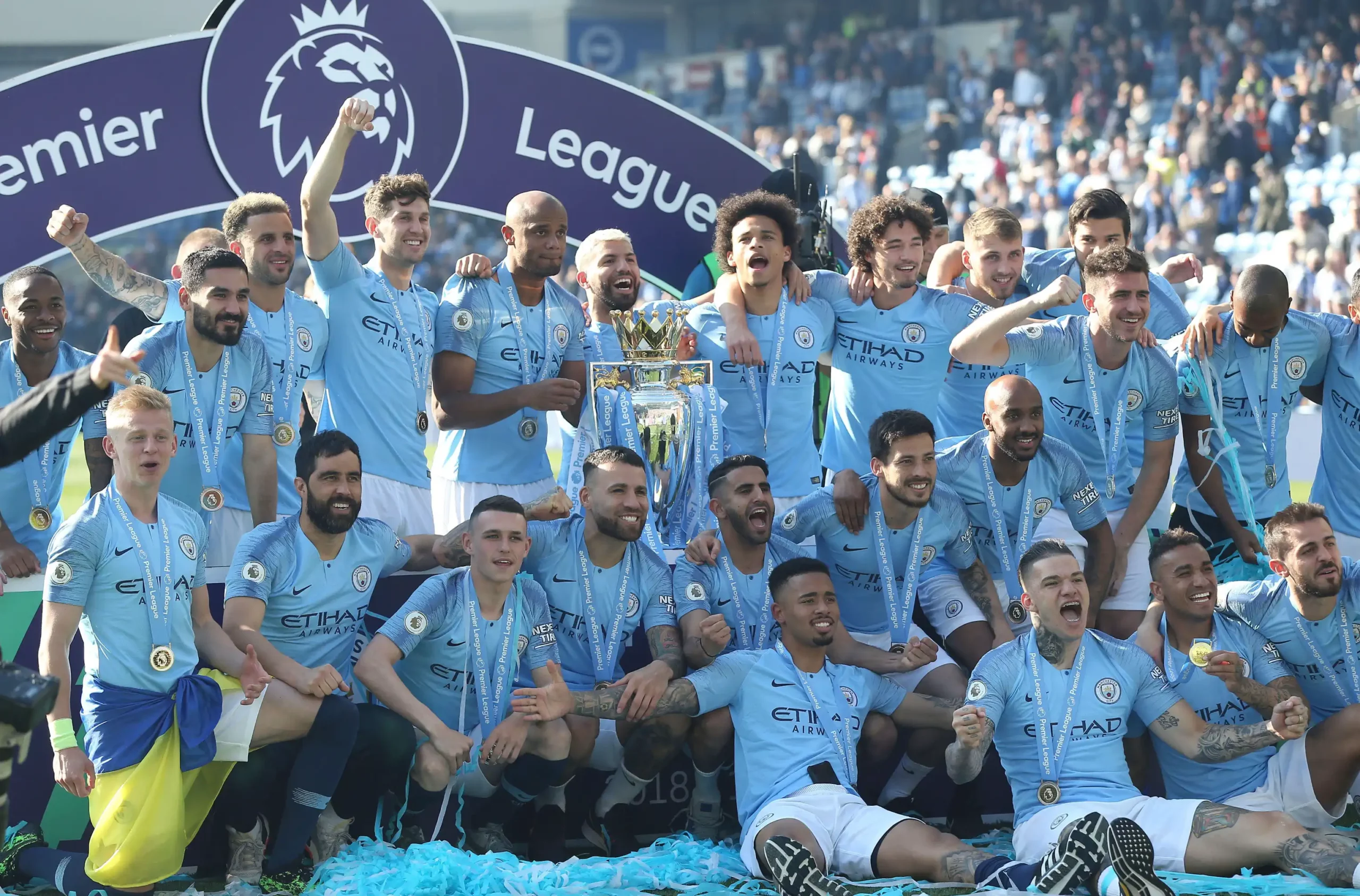A imagem mostra um grupo de jogadores do Manchester City vestindo camisas azuis. Eles estão com os braços levantados em comemoração, enquanto alguns seguram um troféu ou uma taça.