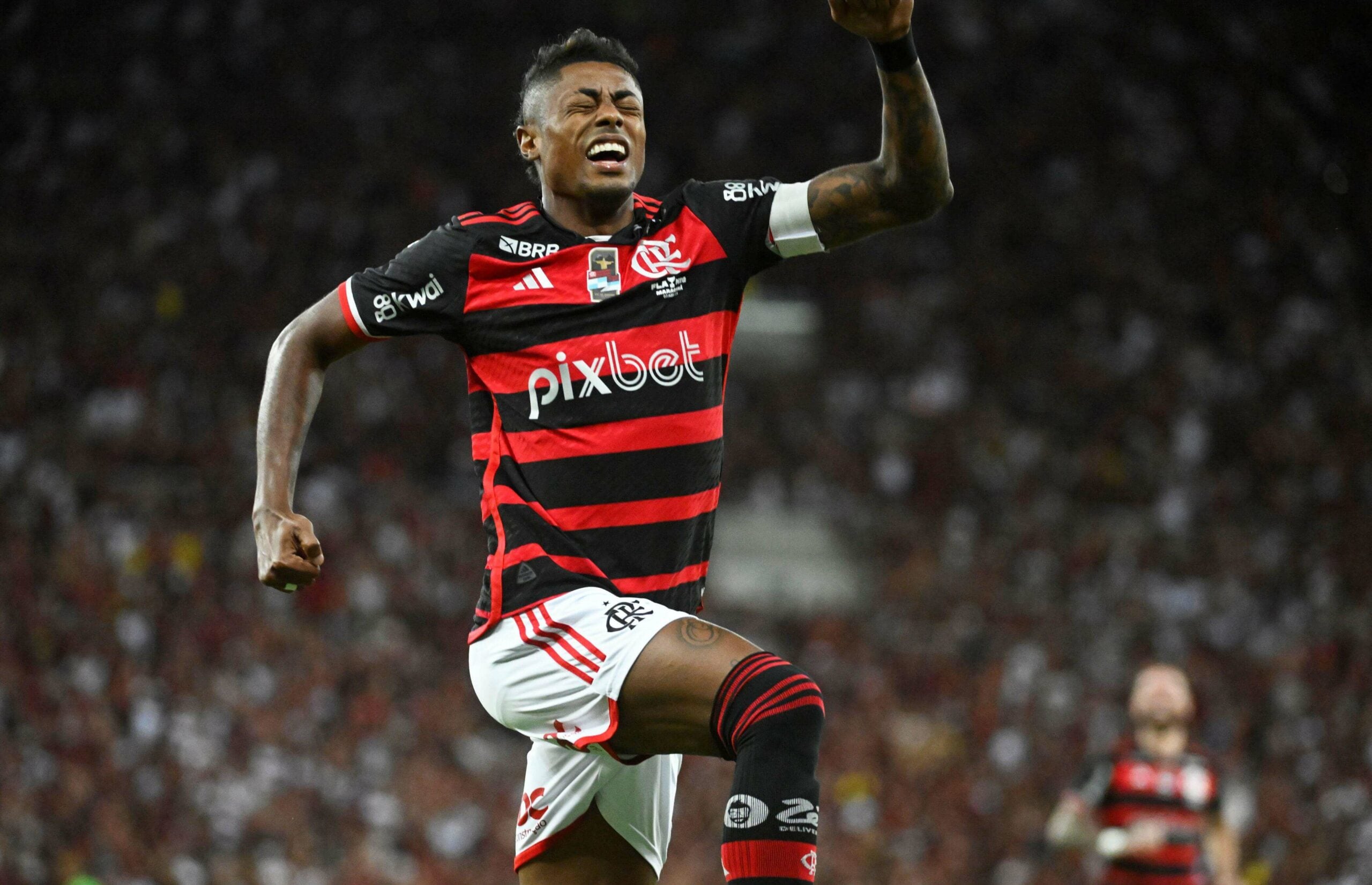 A imagem mostra Bruno Henrique, jogador do Flamengo, comemorando um gol na final do Campeonato Carioca. Ele está com os braços levantados no ar, vestindo uma camisa listrada nas cores vermelha e preta.