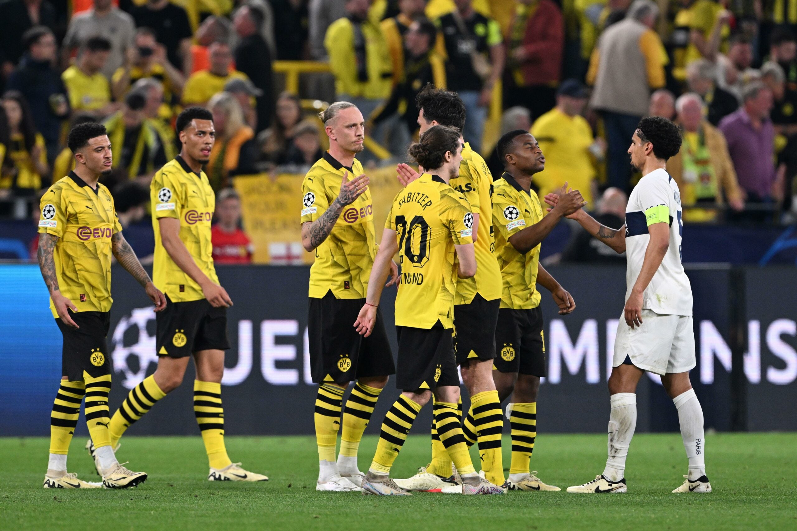 A imagem mostra um grupo de jogadores do Borussia Dortmund vestindo uniformes amarelos cumprimentando jogadores do PSG vestindo uniformes brancos. Eles estão em pé em um campo, com uma arquibancada ao fundo.