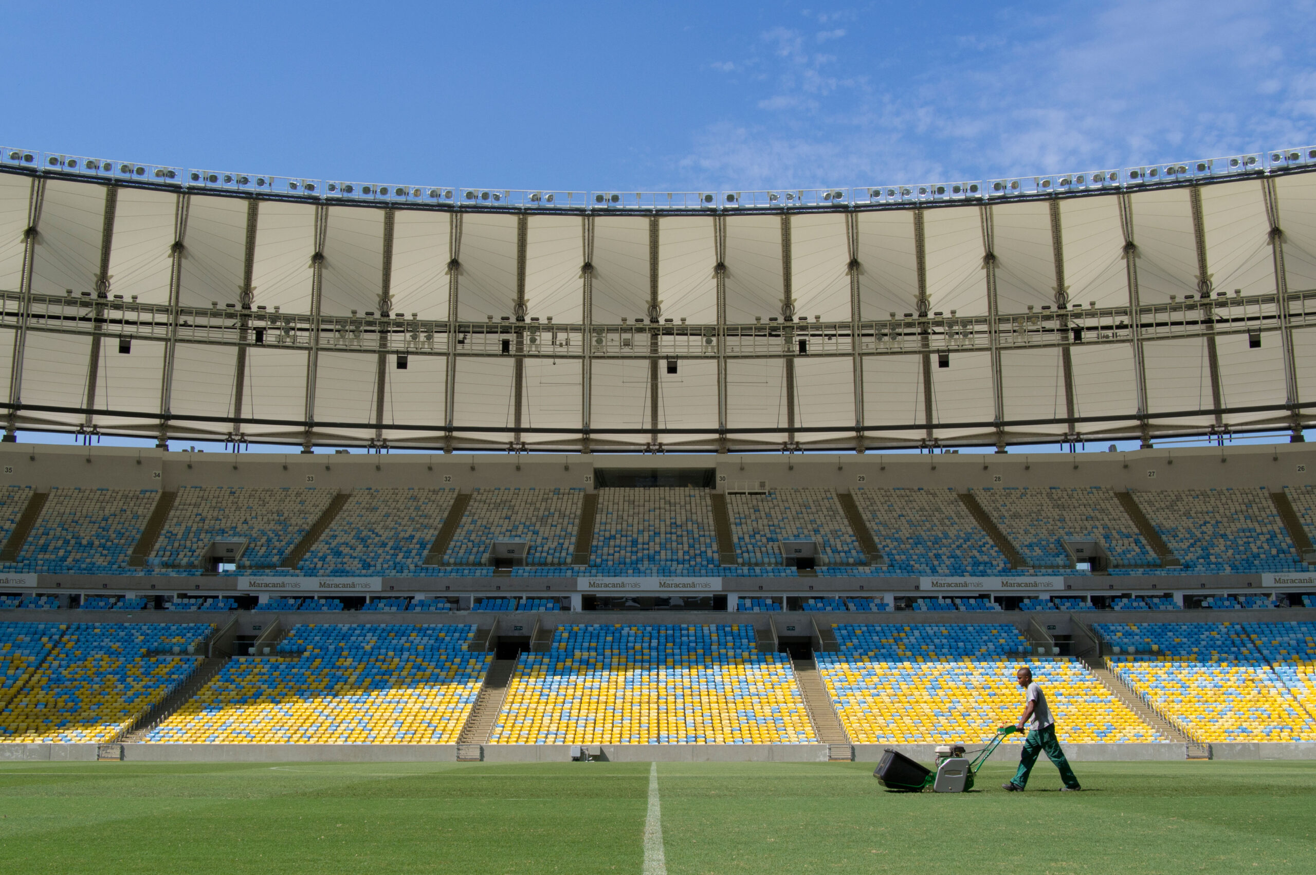 A imagem mostra um funcionário do campo usando um uniforme de limpeza, preparando o gramado de um estádio vazio. Detrás dele está uma arquibancada vazia.