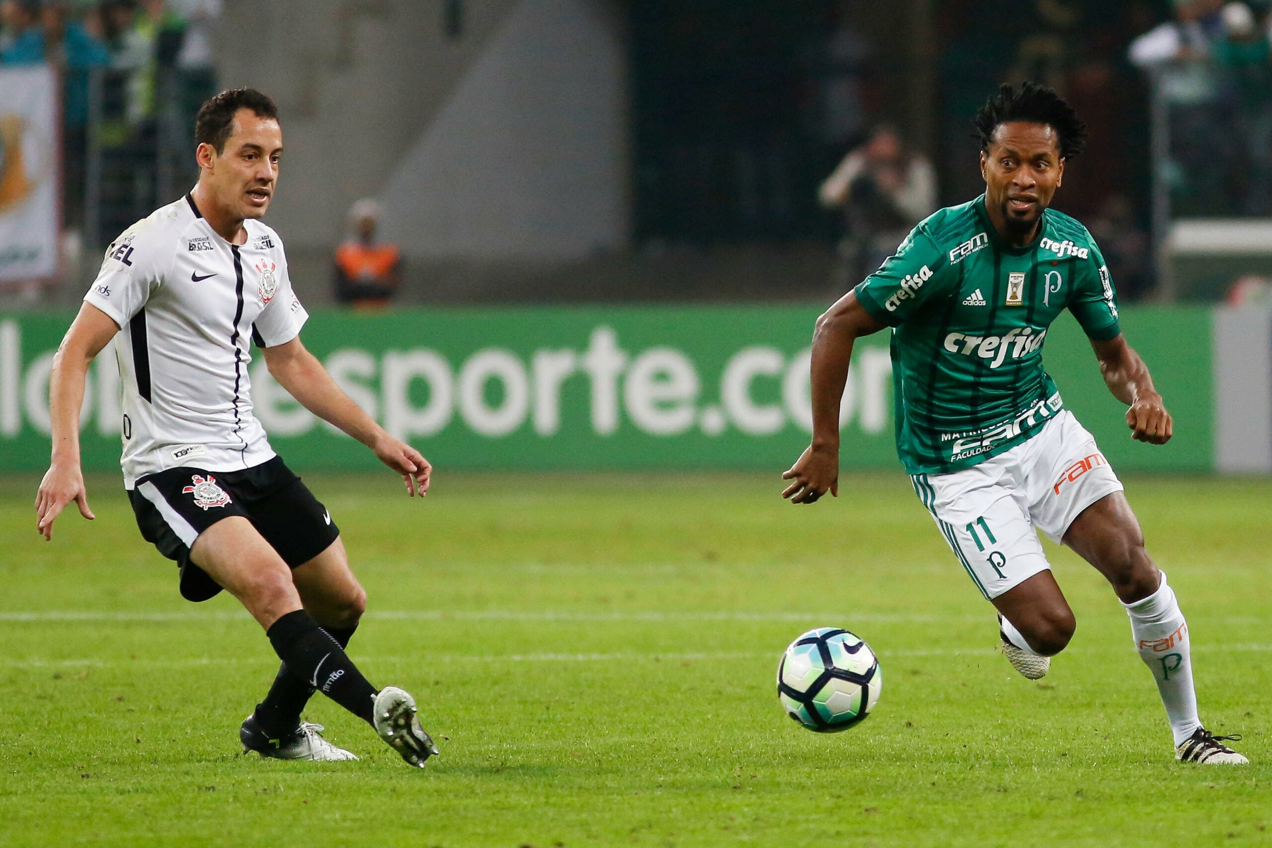 Zé Roberto e Rodriguinho durante a partida entre Palmeiras e Corinthians realizada no Allianz Parque, Zona Oeste de São Paulo. Os jogadores usam o uniforme de seus respectivos times, um na cor verde e outro, branco e preto. Ambos estão disputando pela posse da bola em campo.