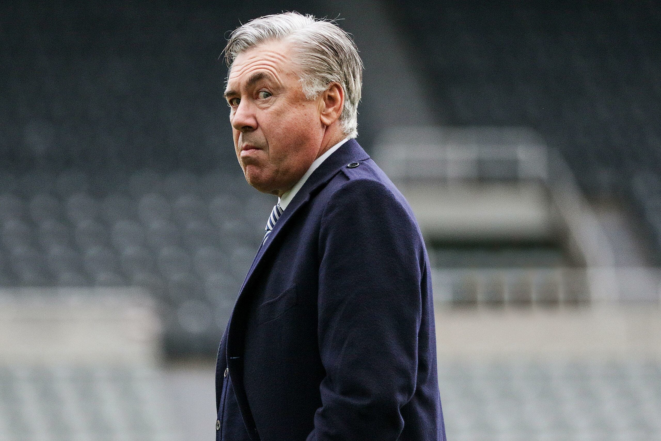 Carlo Ancelotti, treinador italiano, é capturado em um momento reflexivo durante uma partida, vestindo um elegante casaco escuro.