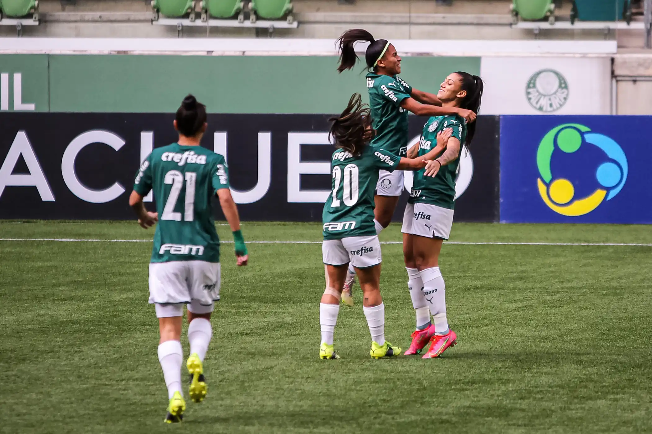 A imagem mostra um grupo de jogadoras do Palmeiras vestindo uniformes verdes comemorando em campo. Elas estão abraçadas umas às outras e exibem expressões de alegria e entusiasmo.