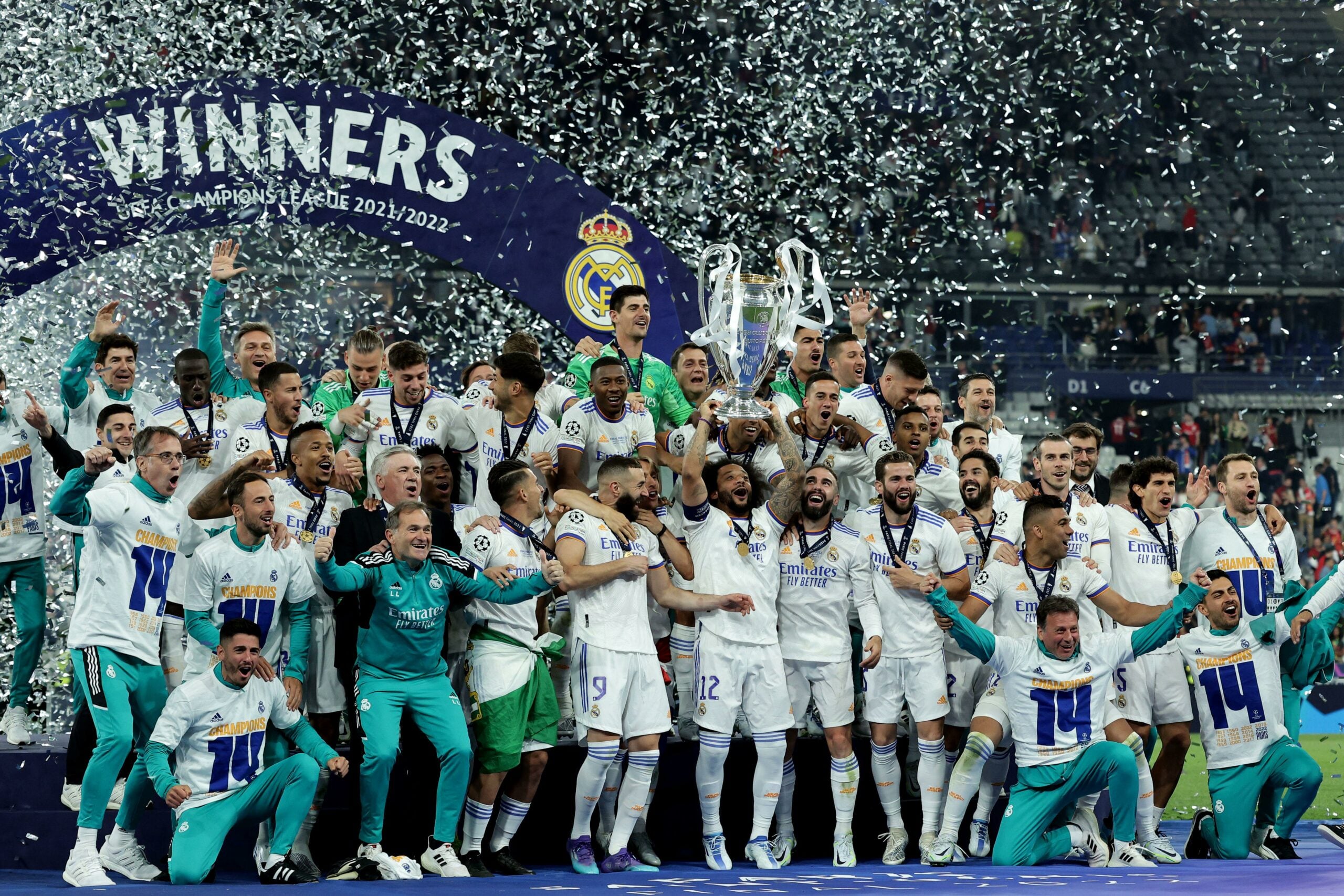 Real Madrid celebrando a conquista do título da Liga dos Campeões 2021/2022. Os membros da equipe seguram um troféu e estão em uma fila, com alguns deles levantando o troféu acima de suas cabeças. O fundo da imagem corresponde a um estádio com alguns espectadores visíveis nas arquibancadas.