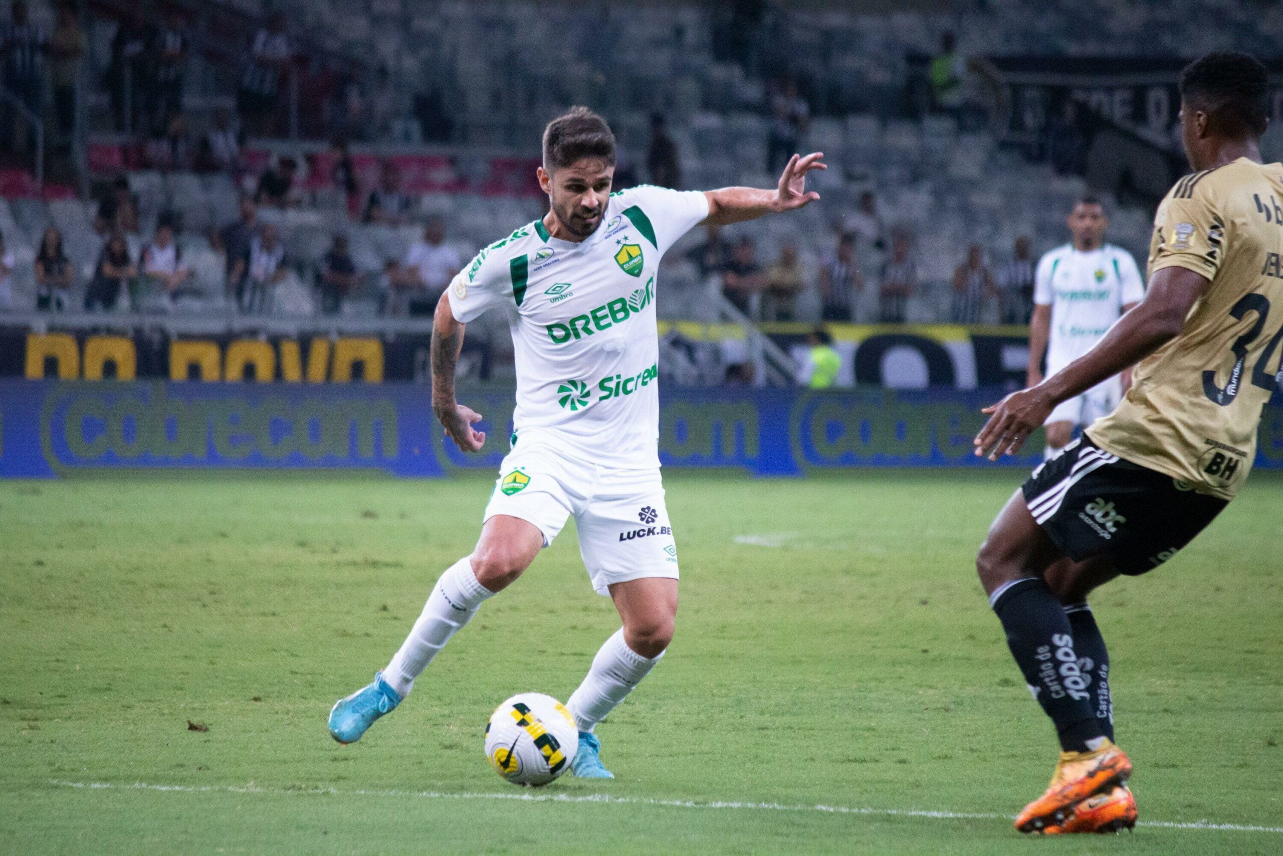 Partida de futebol entre Atlético-MG e Cuiabá, válida pela 37ª rodada do Campeonato Brasileiro de Futebol, realizada no estádio Mineirão, em Belo Horizonte. O jogador do Cuiabá domina a bola e está prestes a chutá-la. Ele usa uniforme branco com detalhes verdes. Ao fundo vê-se a arquibancada. Ao lado dele, há o jogador do Atlético-MG.