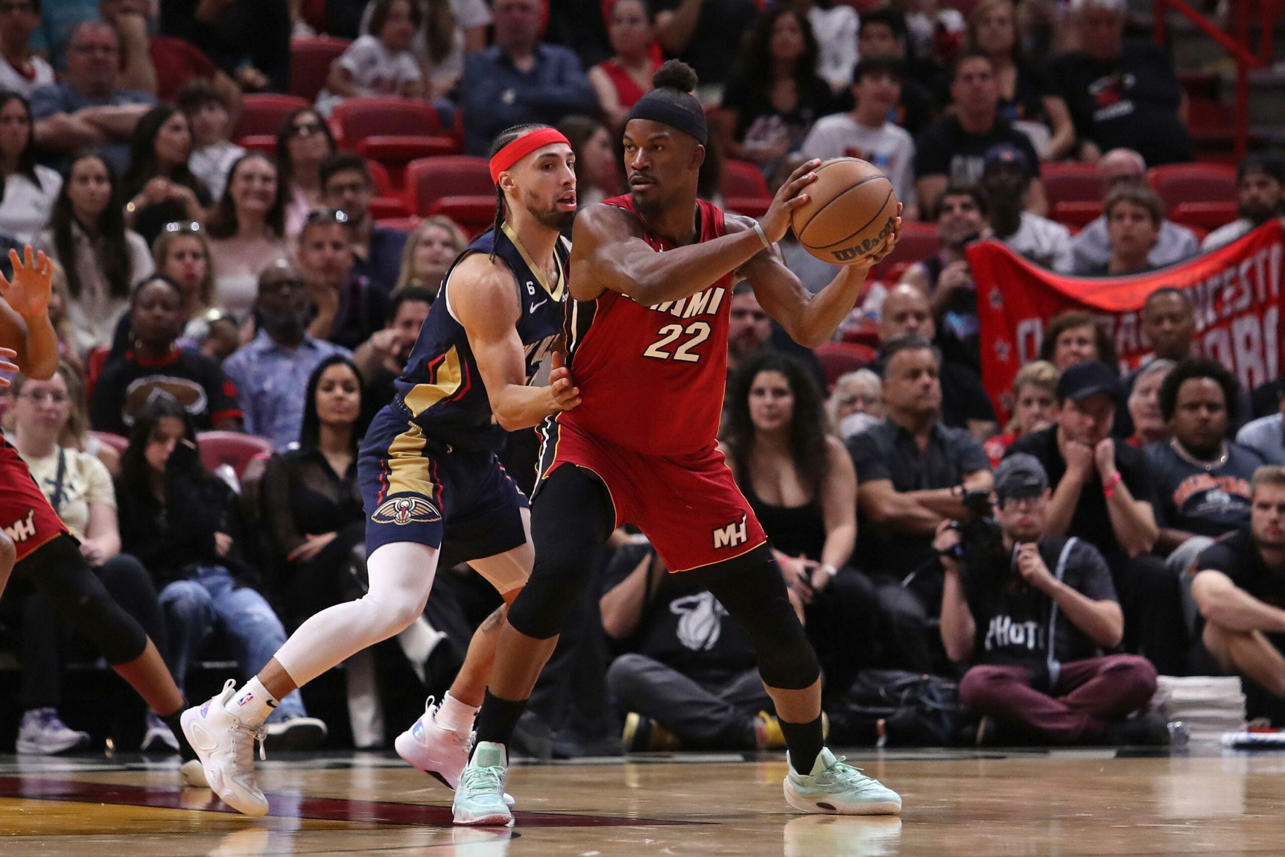 Jimmy Butler, do Miami Heat, vestindo o uniforme vermelho número 22, segura a bola de basquete enquanto é marcado de perto por um jogador do New Orleans Pelicans, com a torcida ao fundo observando atentamente a ação em quadra