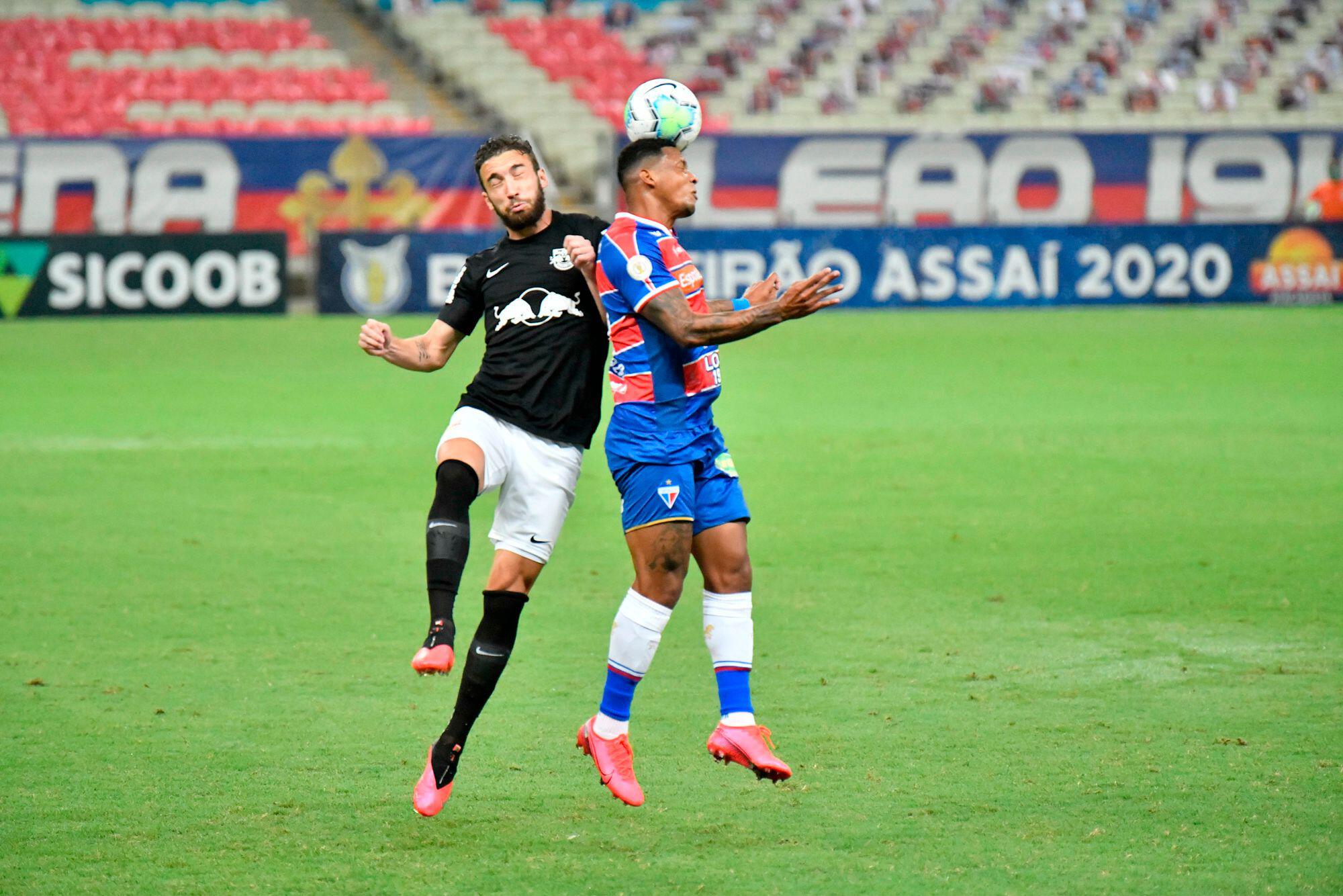 A imagem mostra dois jogadores, um do Fortaleza e outro do Red Bull Bragantino, em um momento de disputa pela posse de bola. Ambos estão vestindo seus respectivos uniformes de seus times. Eles estão em um campo de futebol, com a bola posicionada na cabeça do jogador.