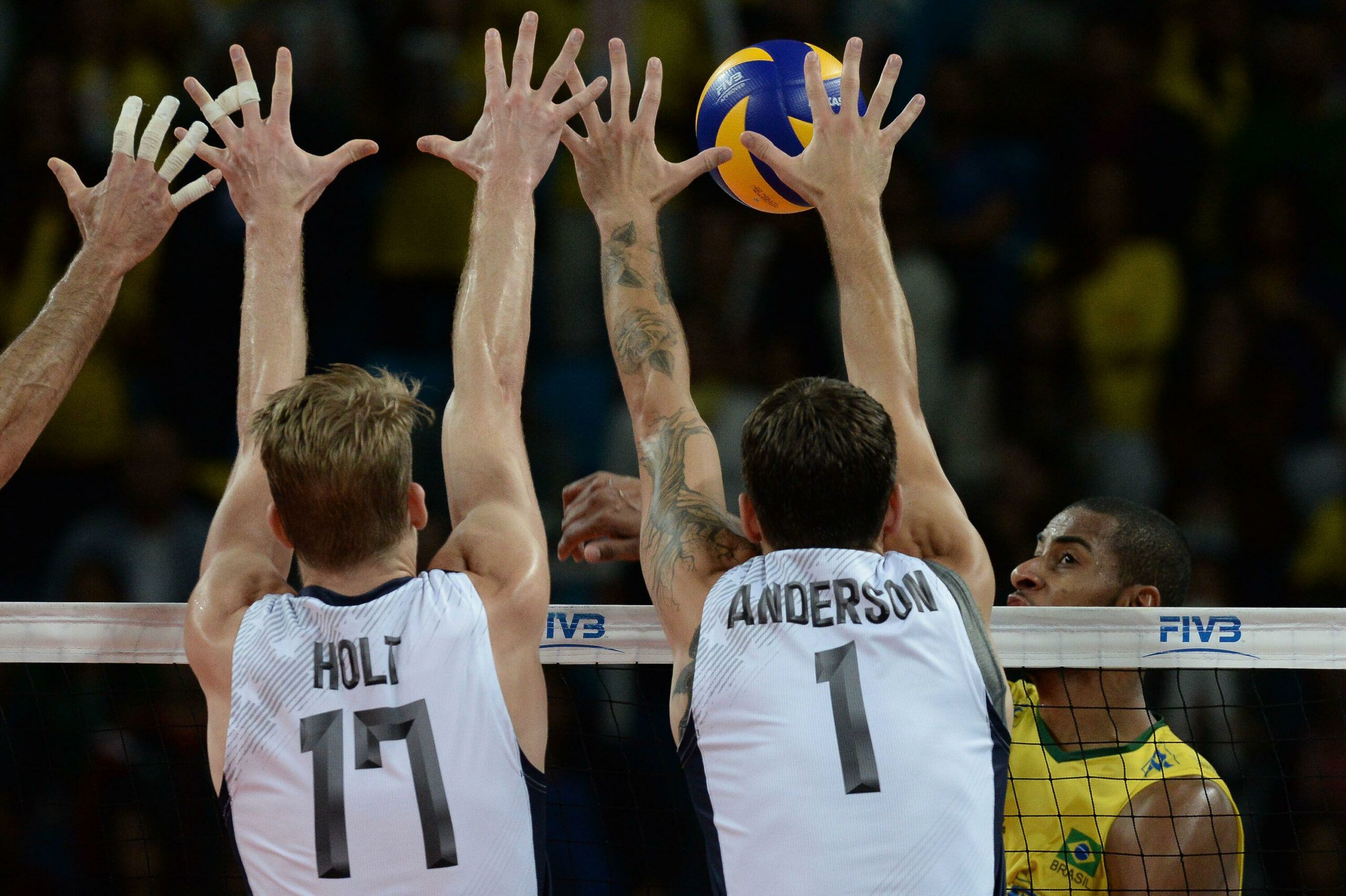 A imagem mostra Lucarelli, jogador da seleção brasileira de vôlei, em pleno jogo contra jogadores dos Estados Unidos. Ele está posicionado para atacar a bola, enquanto os adversários se preparam para bloquear.