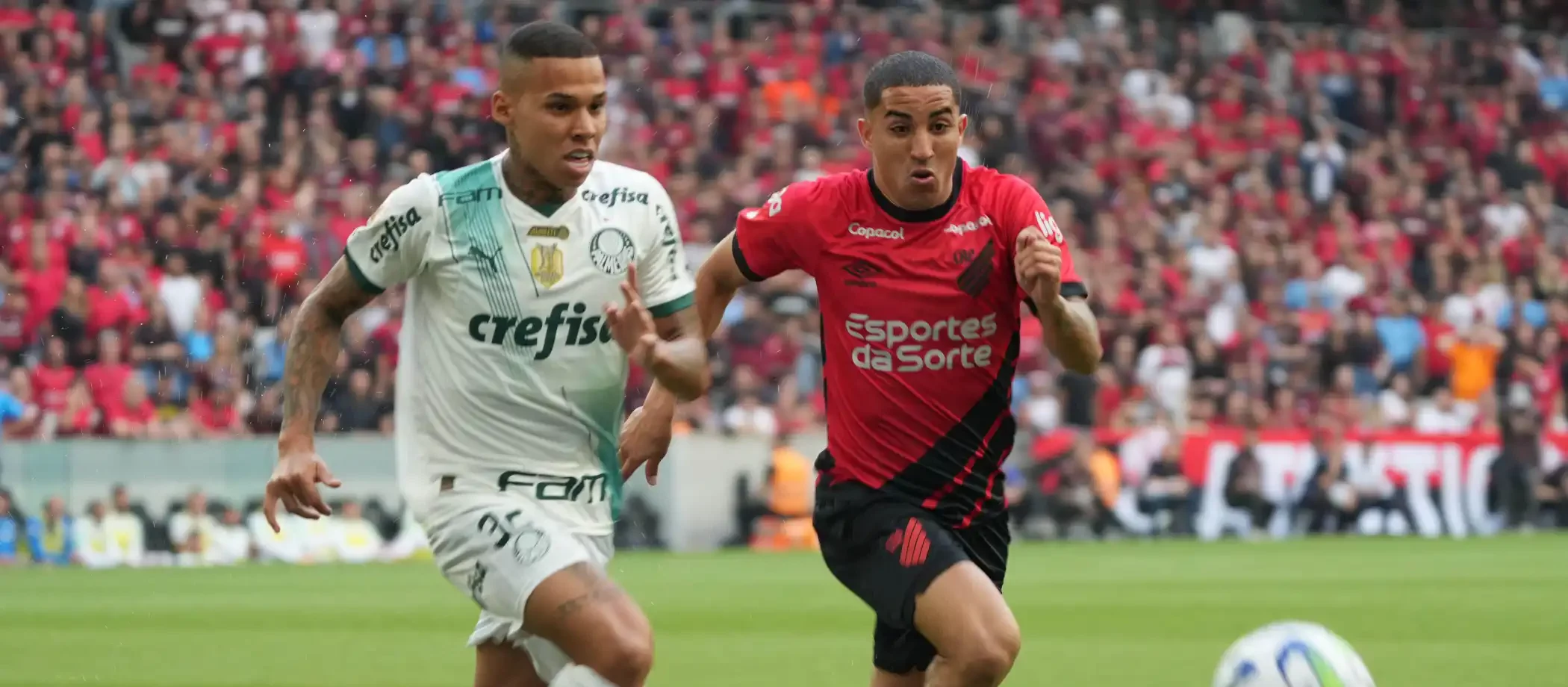 Dois jogadores de futebol, Christian e Garcia, correndo atrás da bola em um campo durante partida entre Athletico-PR e Palmeiras pelo Brasileirão.