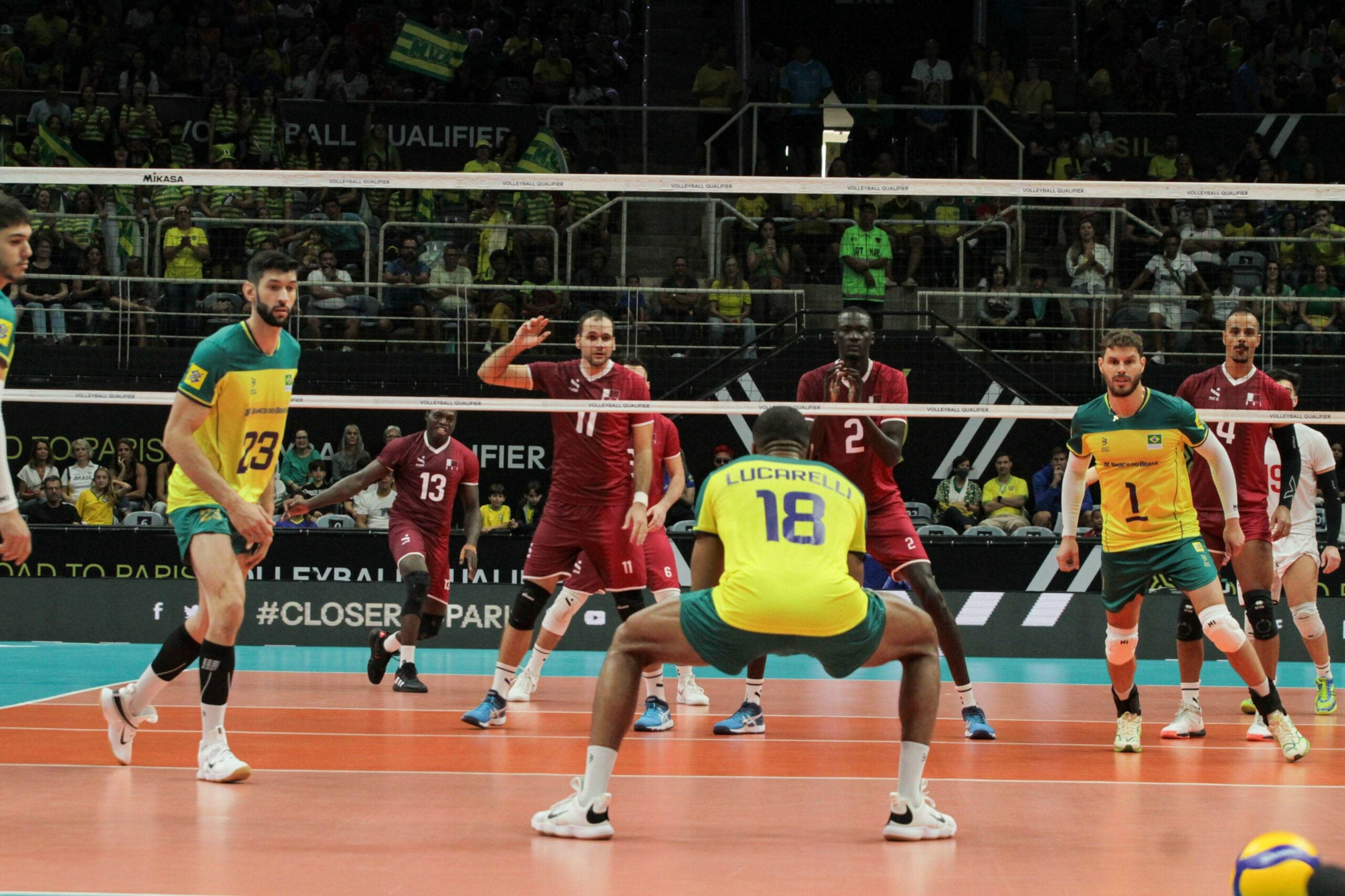 Em quadra, a seleção brasileira de vôlei enfrentava o Qatar pelo Pré-olímpico de Vôlei Masculino em 2023. A equipe veste uniforme amarelo e verde, em contraste com o adversário, que veste uniforme vermelho. O jogador Lucarelli se prepara para defender a bola