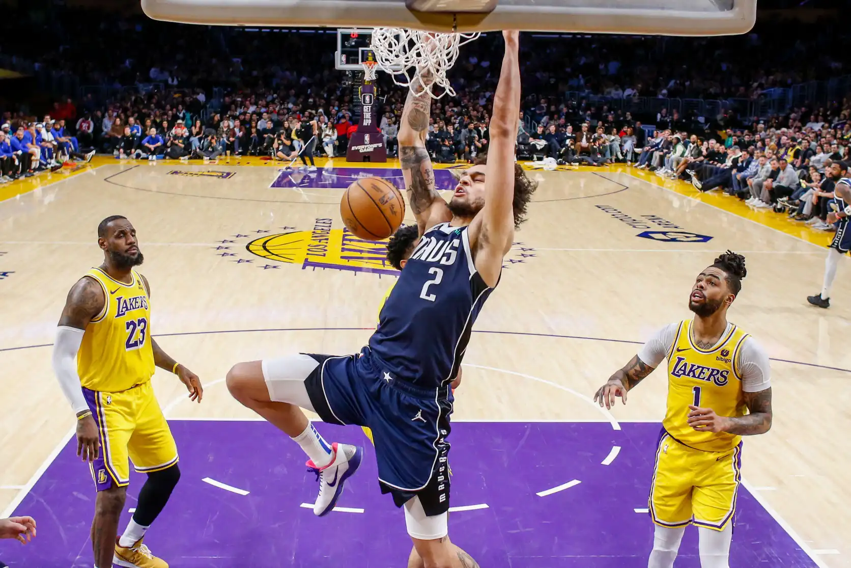 Dereck Lively II, jogador do Dallas Mavericks, está no meio de um salto, estendendo o braço para enterrar a bola na cesta. O jogador usa uniforme amarelo e há dois jogadores do time adversário, Lakers, ao lado dele.