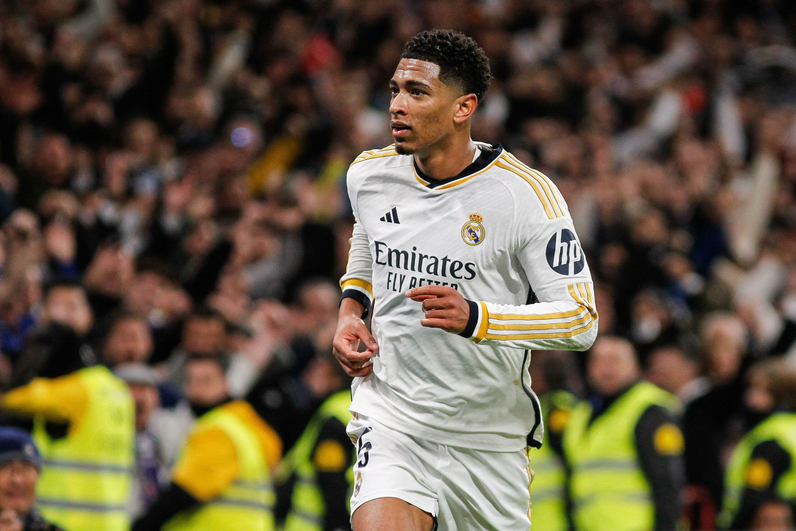 A imagem mostra Jude Bellingham, jogador do Real Madrid, correndo em campo com a bola. Ele está vestindo um uniforme branco do Real Madrid, com o logotipo do time na frente. Atrás dele, há uma arquibancada com torcedores.