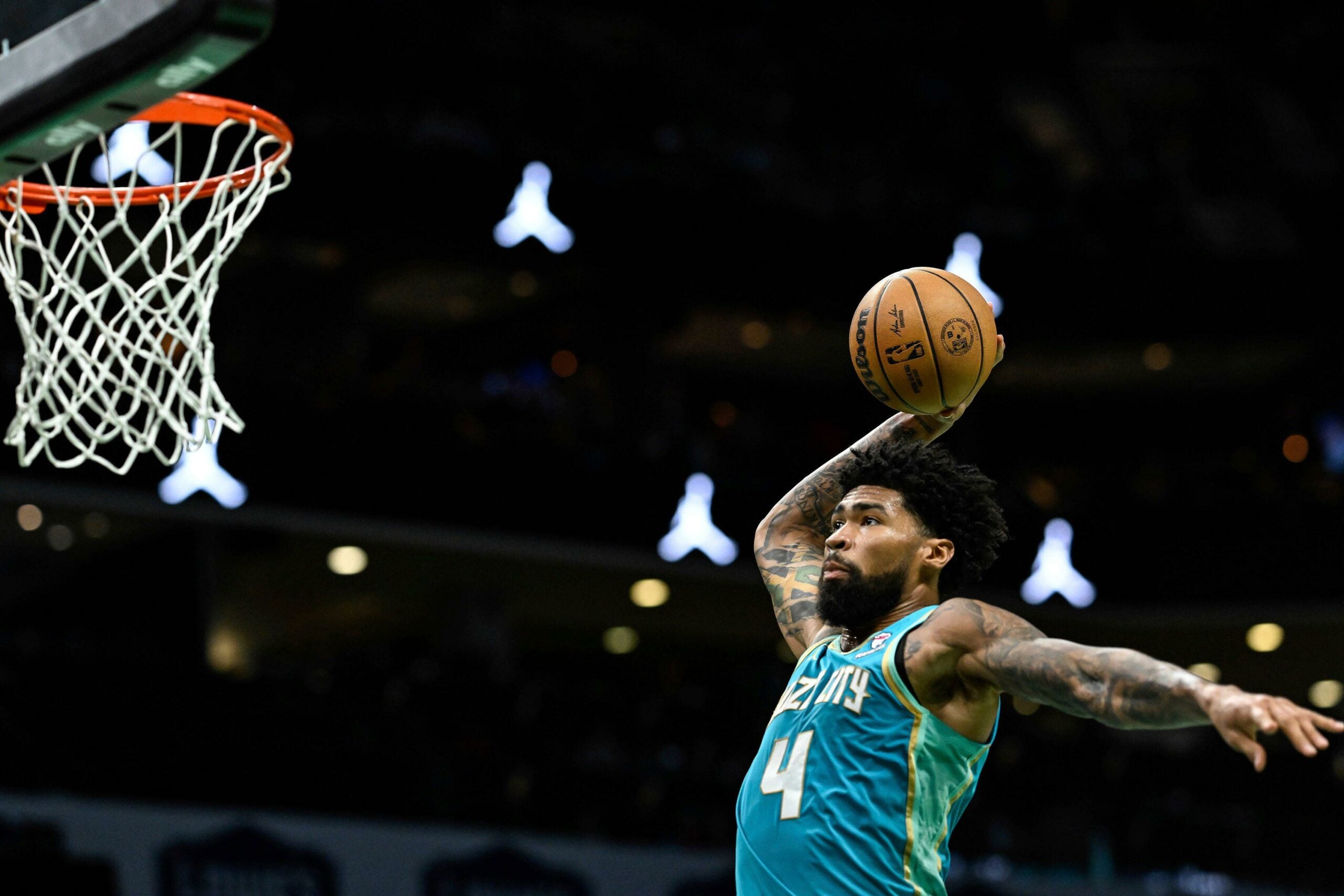 Jogador do Charlotte Hornets em uniforme turquesa se prepara para uma enterrada, segurando a bola de basquete com uma mão, enquanto se eleva próximo à cesta durante um jogo, destacando seus braços tatuados.