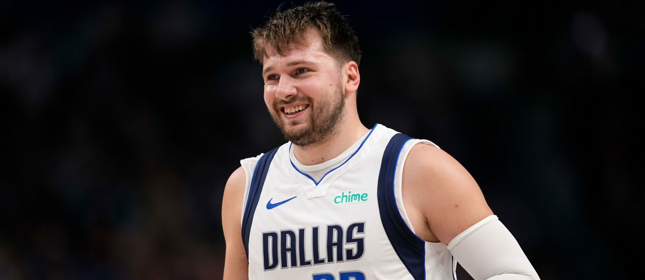 Luka Dončić, do Dallas Mavericks, cotado para MVP da temporada 23/24, sorri durante jogo da NBA. Ele usa uniforme branco com detalhes em azul.