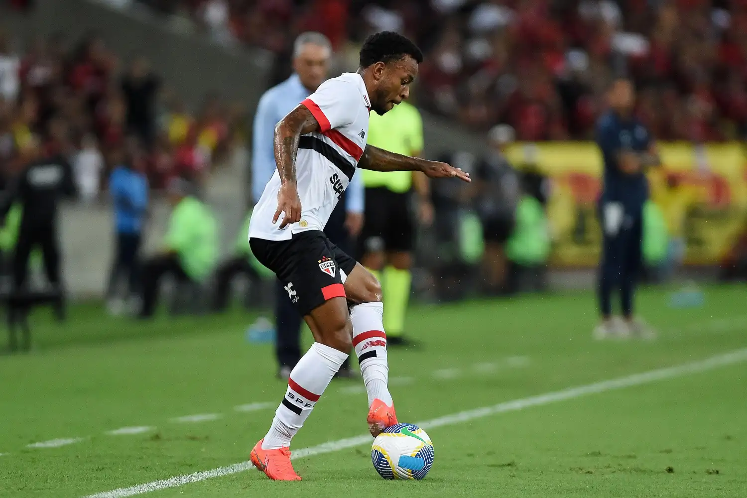 Jogador do São Paulo domina bola em campo. Usa uniforme branco com listras vermelha e preta. O técnico do time está desfocado atrás do jogador.