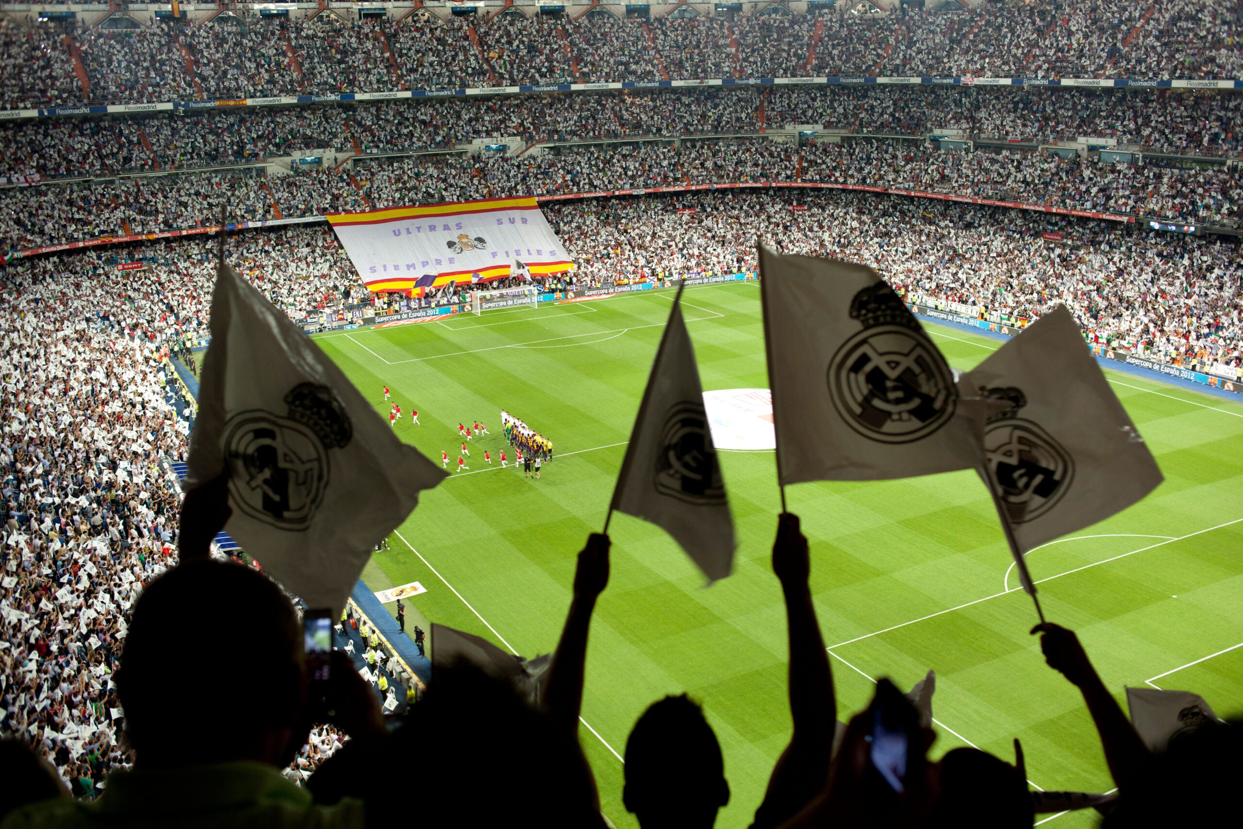 A imagem mostra uma vista panorâmica do Novo Santiago Bernabéu, o estádio do Real Madrid. As arquibancadas estão completamente lotadas de torcedores empolgados, que exibem bandeiras brancas, representando a presença de uma grande multidão.