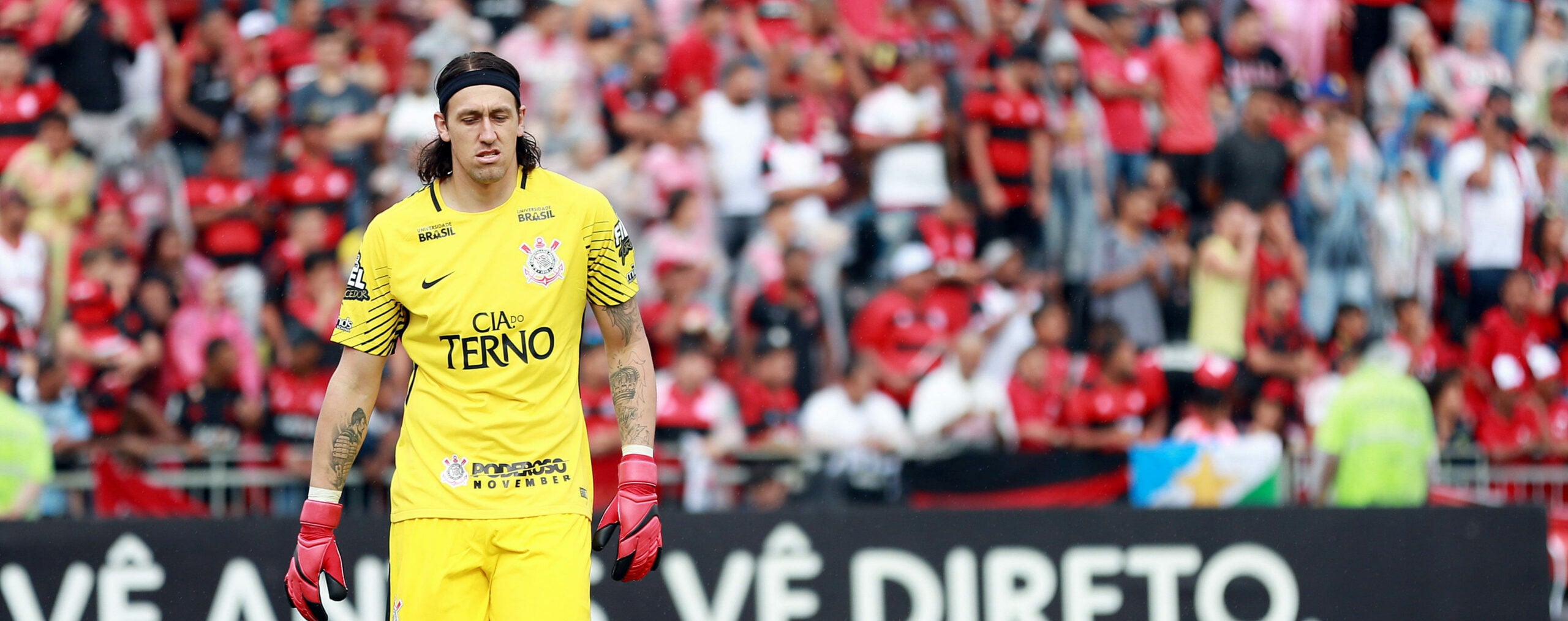 Goleiro Cassio Ramos, do Corinthians, em ação durante a partida contra o Flamengo. Ele está vestindo um uniforme amarelo e luvas vermelhas, e está caminhando pelo campo de futebol.