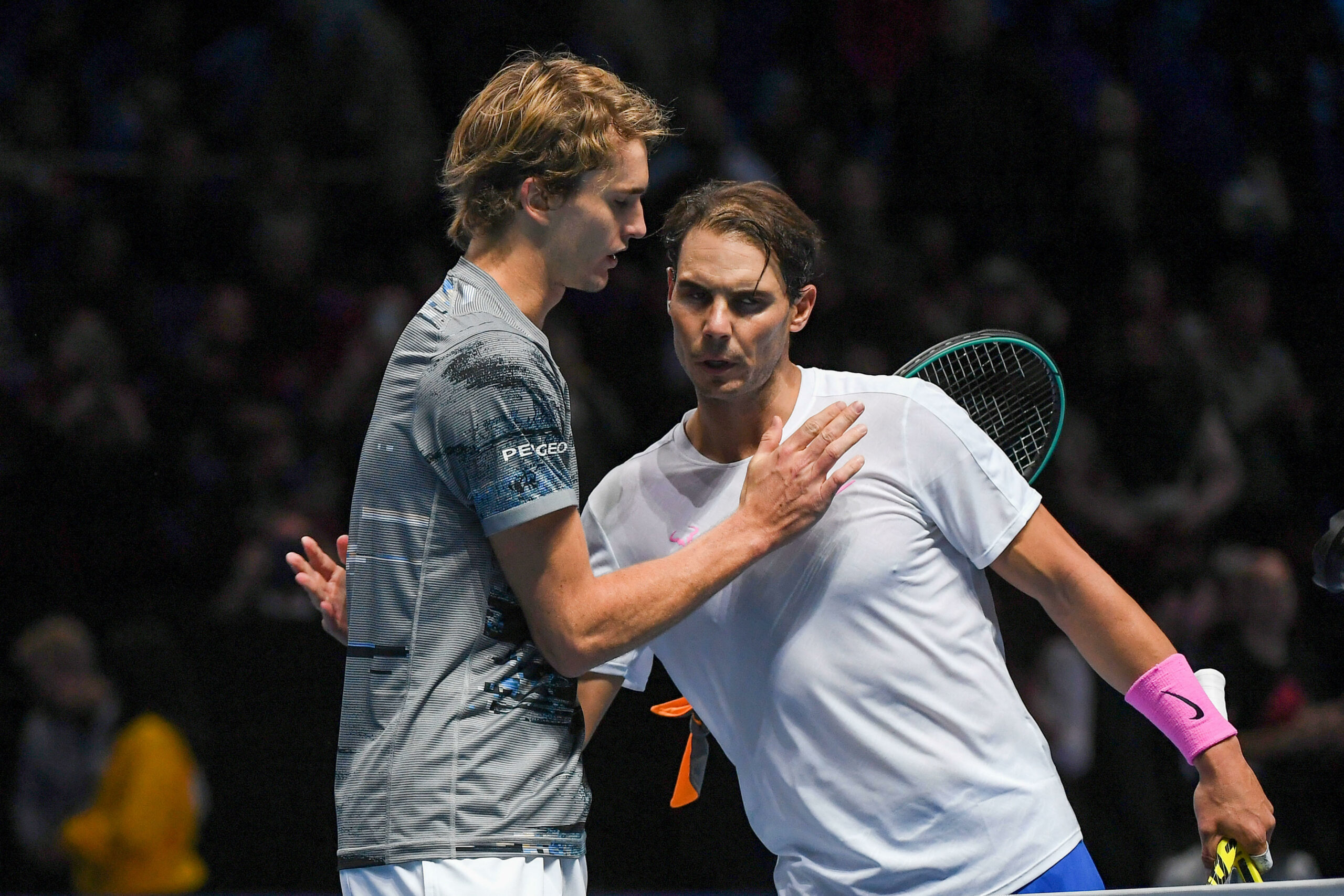 A imagem mostra os jogadores de tênis Rafael Nadal e Alexander Zverev cumprimentando-se no final de uma partida. Ambos estão em pé em uma quadra de tênis, com uma raquete visível ao fundo.