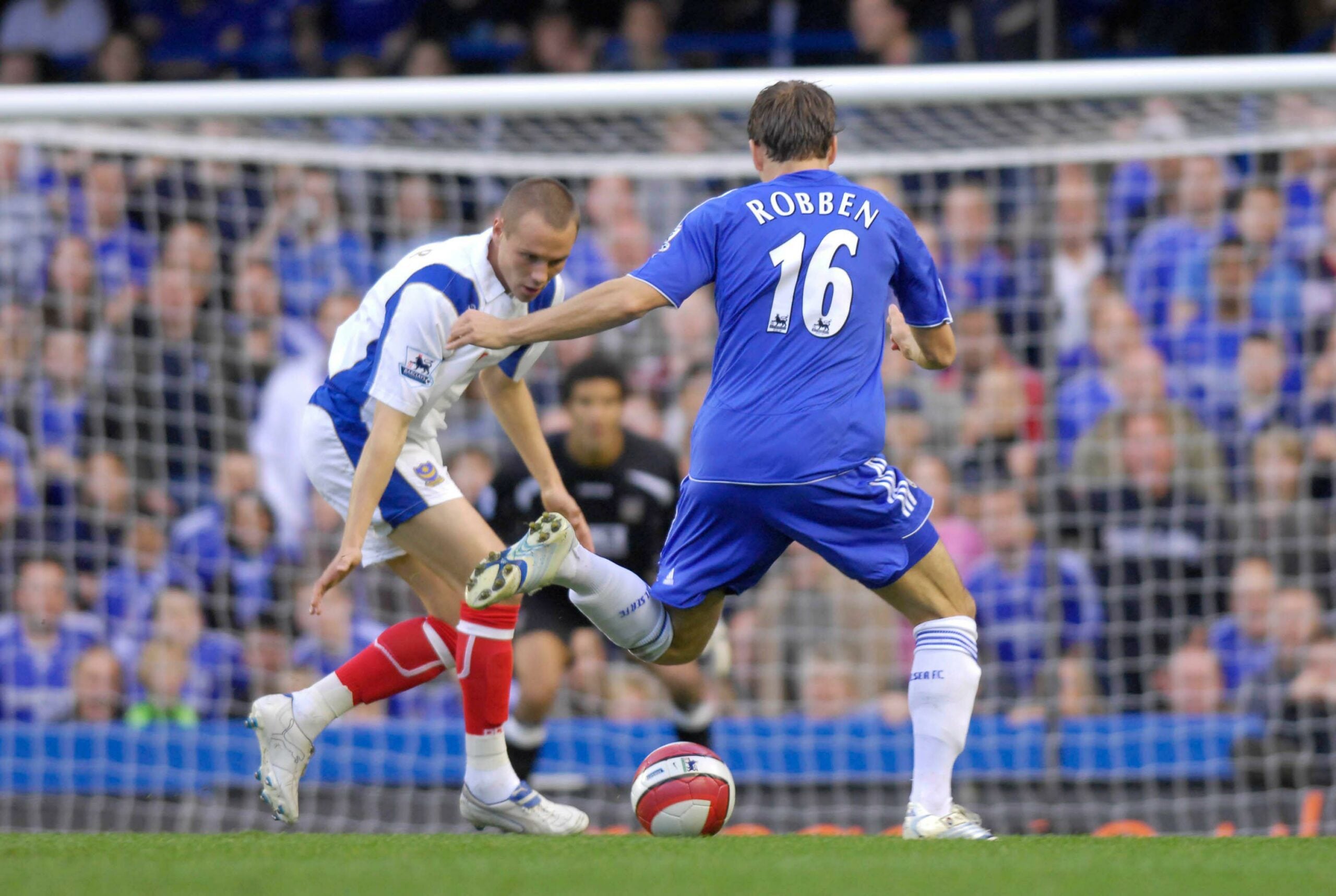 Jogador do Chelsea, Arjen Robben, conduz a bola enquanto é marcado por um defensor do Portsmouth durante uma partida, ambos em ação no campo com o fundo de torcedores nas arquibancadas.