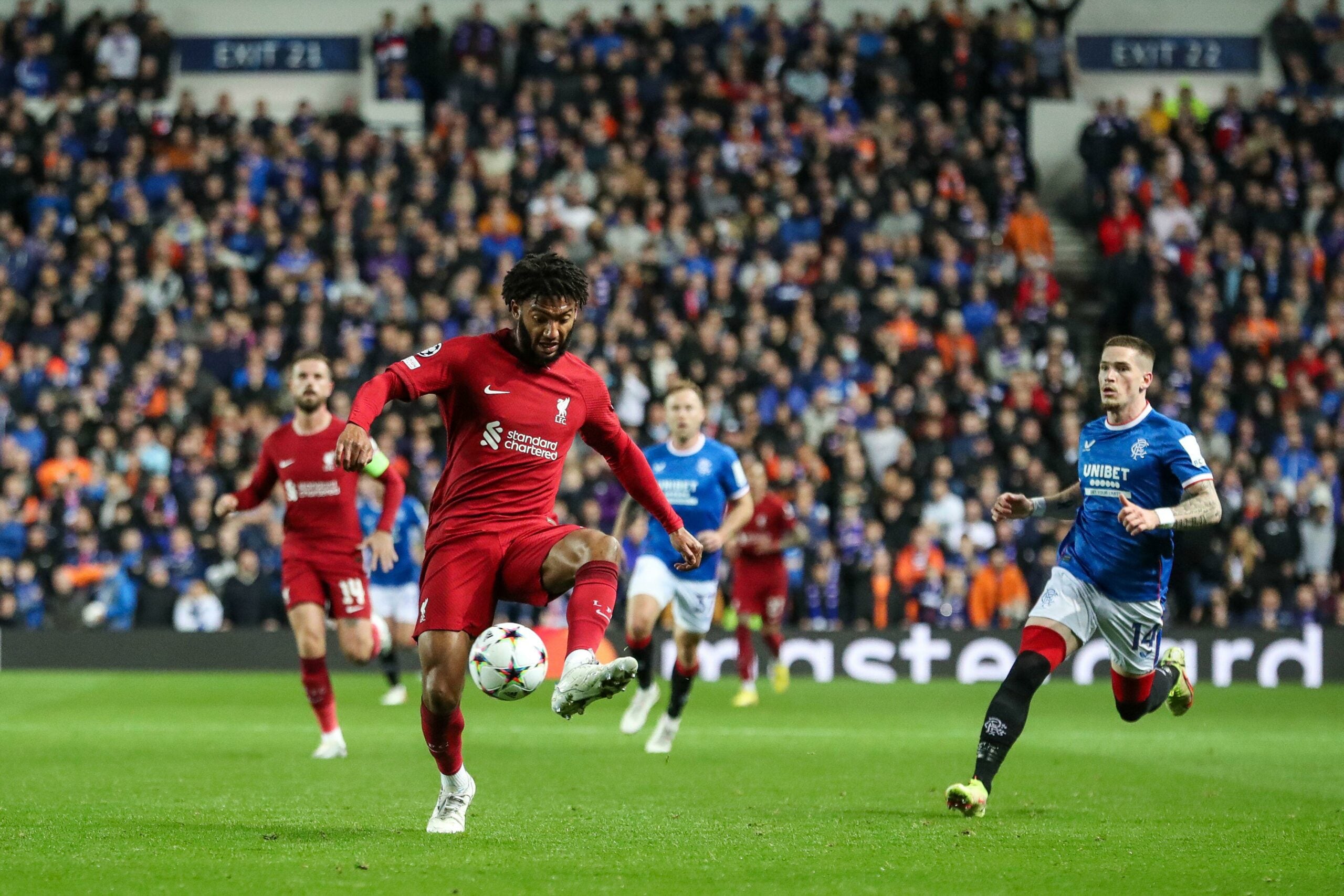 Jogador do Liverpool domina a bola enquanto é perseguido por um defensor do Rangers, durante uma partida com a multidão de torcedores ao fundo, todos em ação no campo.