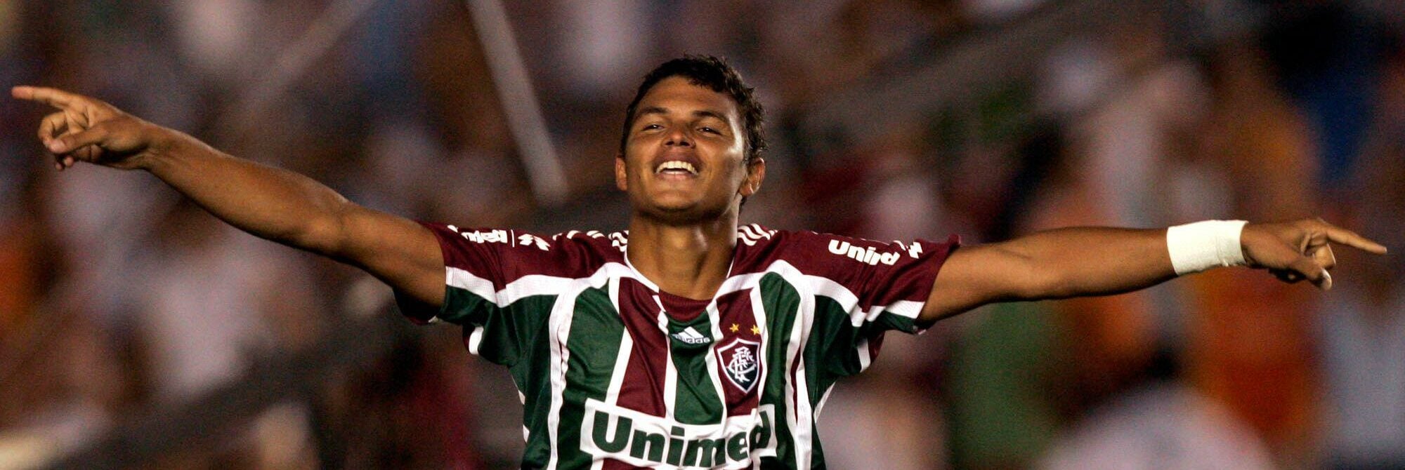O jogador Thiago Silva, do Fluminense, comemora um gol. Ele veste uma camisa verde e branca listrada do time e está levantando o braço direito em celebração. O fundo está desfocado, mas se trata de um estádio cheio de torcedores na arquibancada.