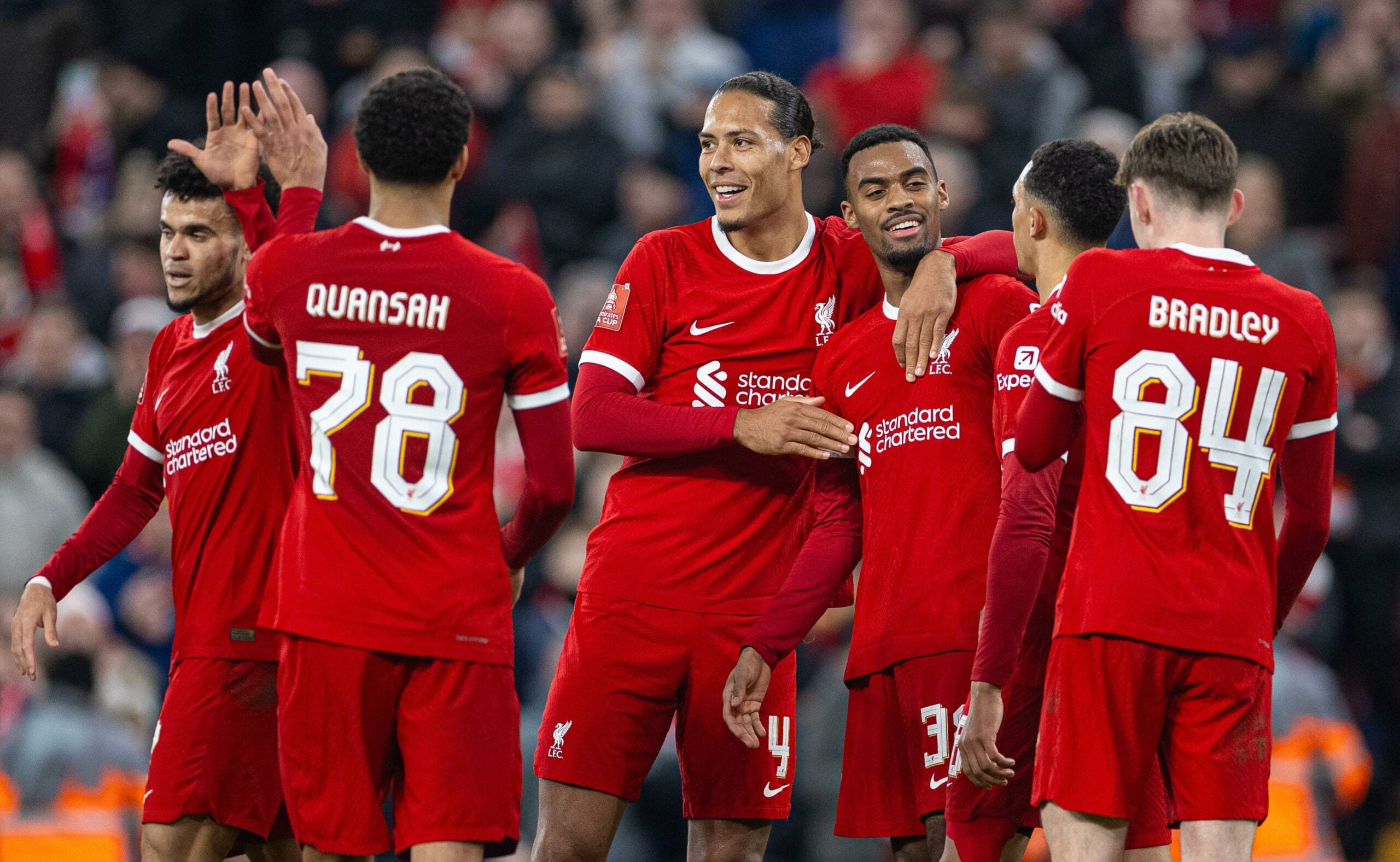 Jogadores do Liverpool, incluindo Virgil van Dijk e Jarell Quansah, comemoram juntos no campo, vestindo o uniforme vermelho, durante uma partida com uma multidão de torcedores ao fundo.