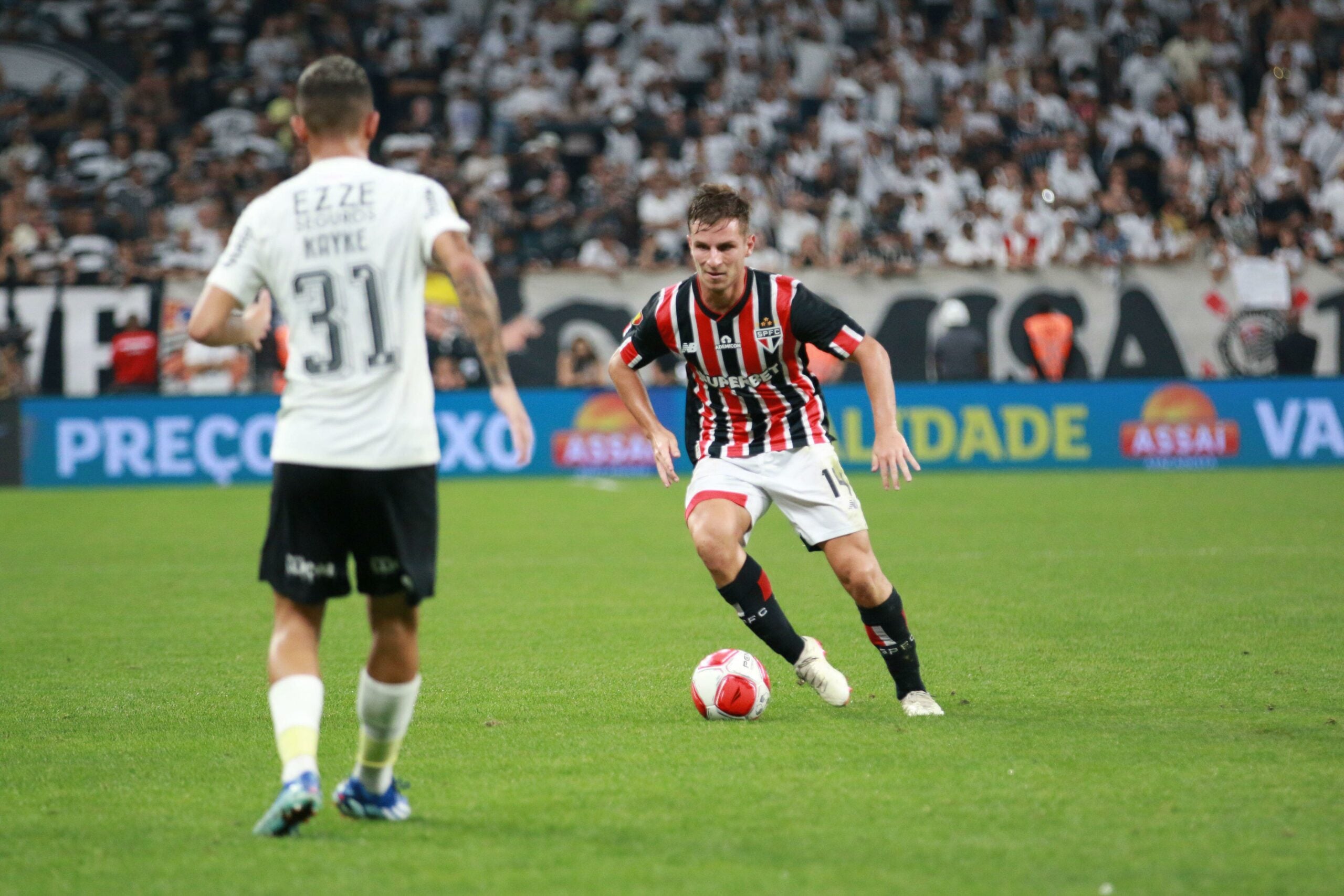Partida de futebol entre Corinthians e São Paulo (Majestoso), válida pela quarta rodada do Campeonato Paulista 2024, realizada no Estádio Neo Química Arena Corinthians, na cidade de São Paulo. O jogador do São Paulo está com a posse da bola e se prepara para chutar. Ele usa o uniforme do time, com listras vermelhas e pretas e detalhes em branco. O jogador do Corinthians se aproxima para fazer a marcação.