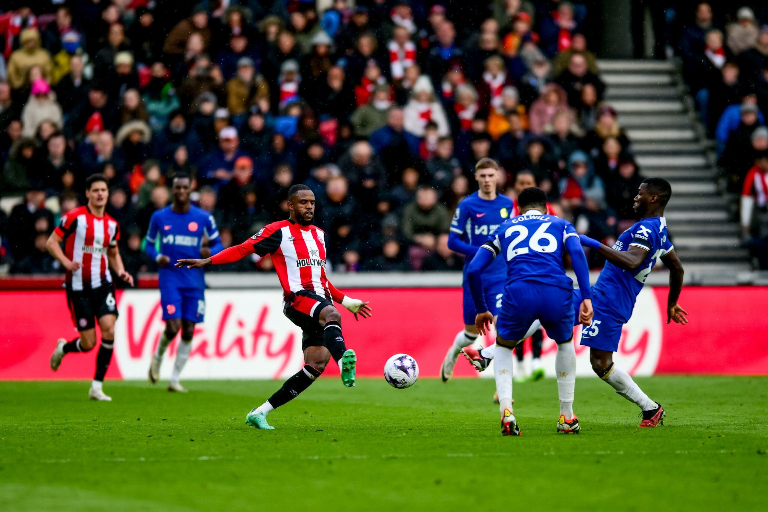 Um jogador do time Brentford com uniforme listrado vermelho e branco disputa a bola com jogadores do Chelsea, que vestem uniformes azuis, durante uma partida de futebol enquanto a torcida assiste atenta.