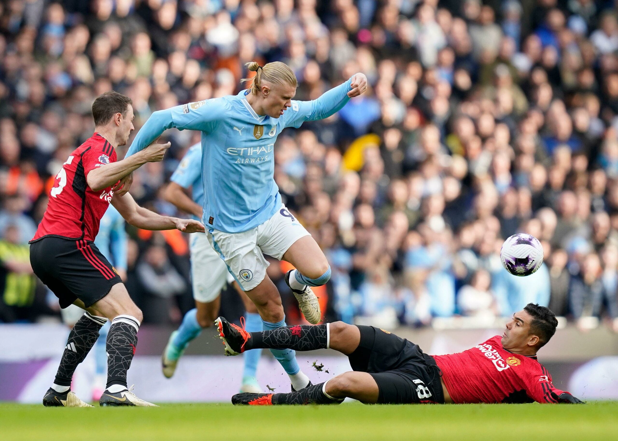 Erling Haaland, do Manchester City, disputa a bola com dois jogadores do Manchester United, um tentando desarmá-lo enquanto o outro está no chão, durante uma partida com uma multidão de torcedores ao fundo.