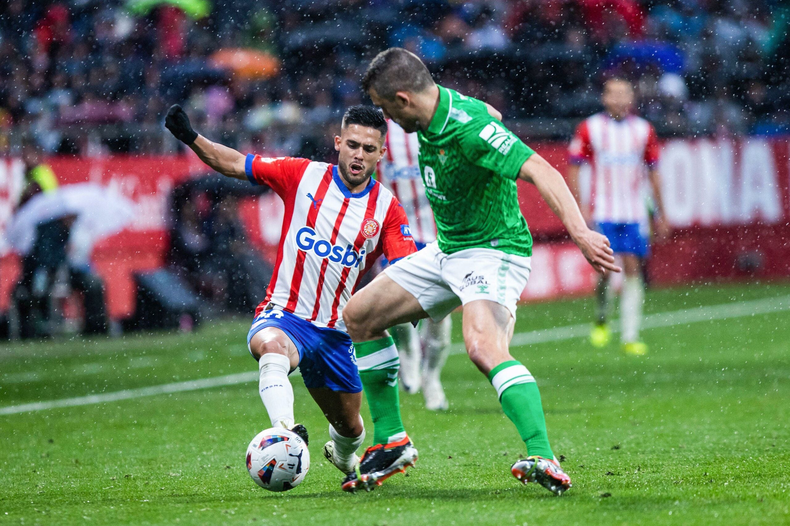 Jogador vestindo a camisa vermelha e branca do Girona, disputando a bola com um oponente de uniforme verde.