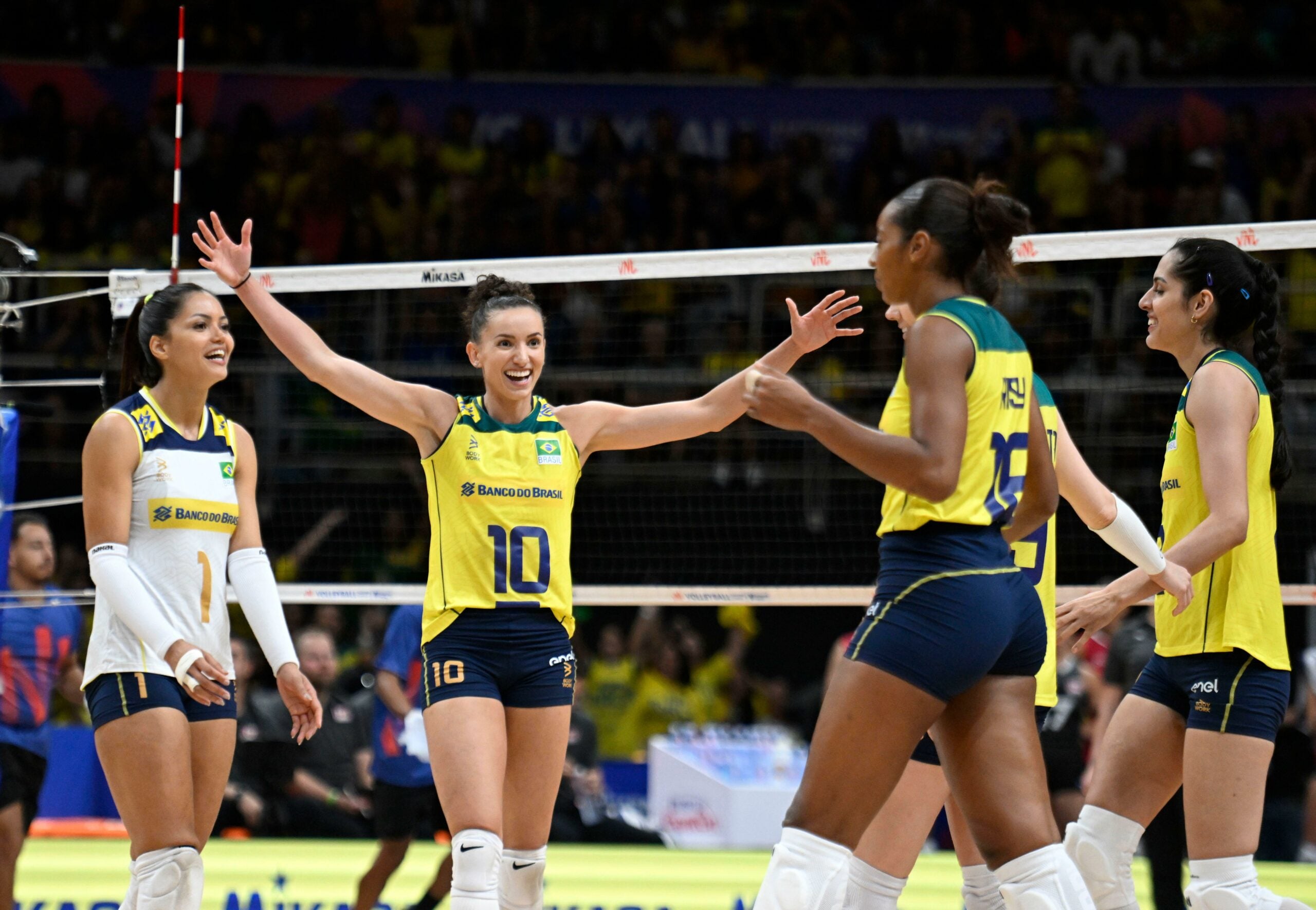 Jogadoras da seleção brasileira de vôlei comemoram ponto em quadra, vestindo uniformes amarelos e azuis, com uma jogadora ao centro, número 10, de braços abertos e sorrindo, enquanto suas companheiras a cercam com expressões de alegria e celebração.