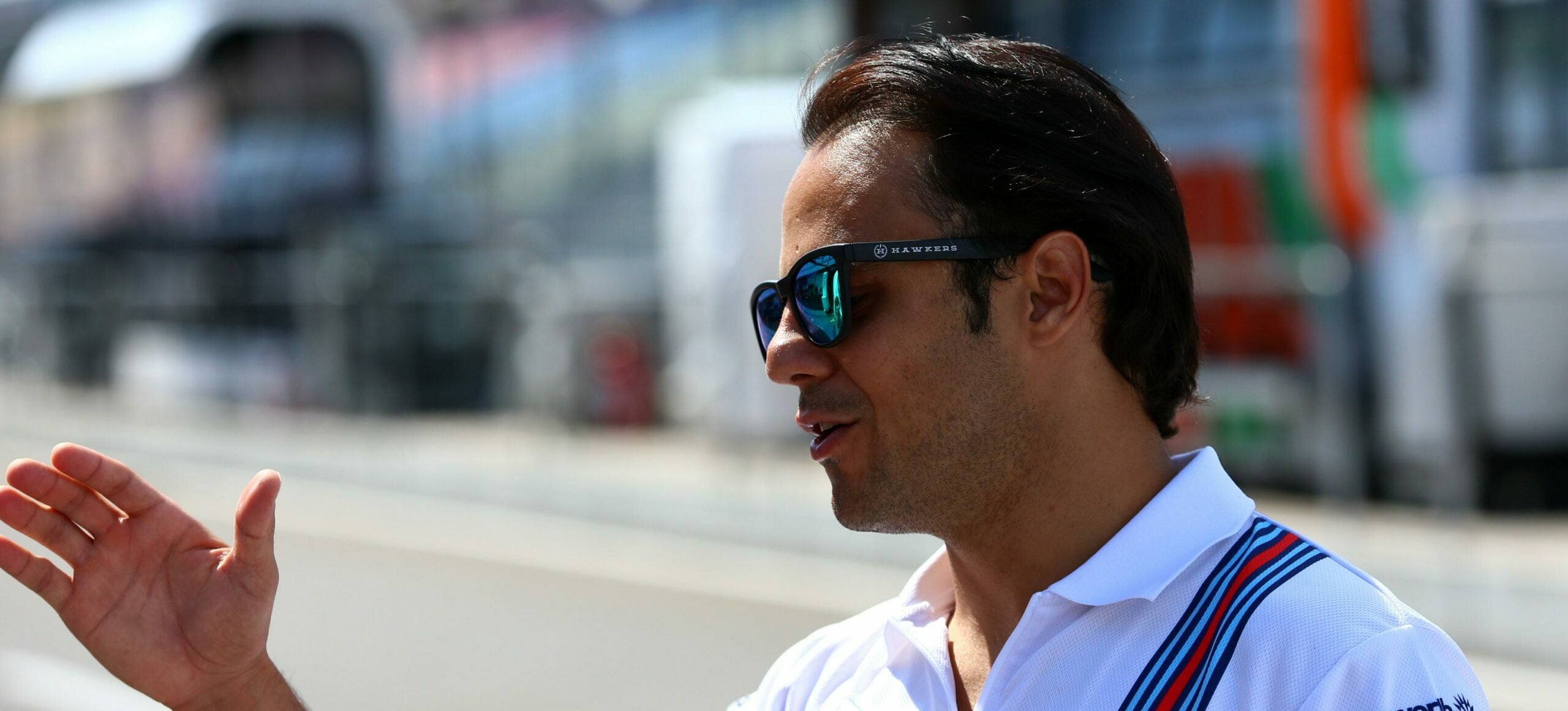 Felipe Massa, piloto brasileiro, veste camisa branca com detalhe em listra azul e vermelha. Ele usa óculos de sol e está na pista de competição da Fórmula 1. O piloto está cumprimentando alguém com a mão direita.