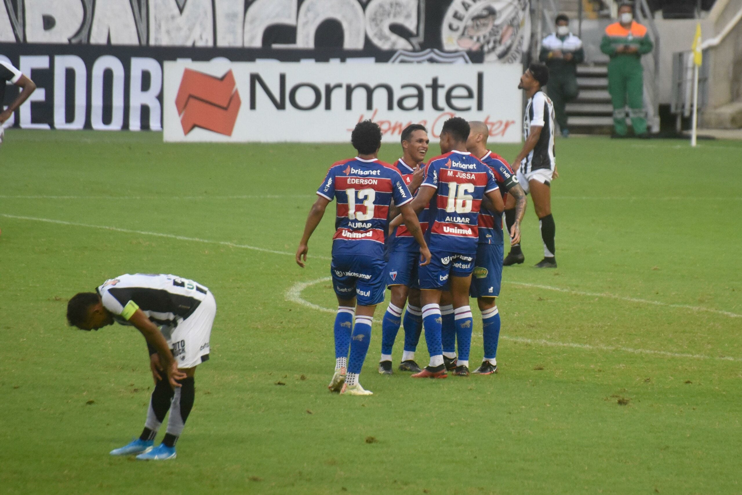 Na imagem, podemos ver um jogador de futebol vestindo uniforme preto, aparentemente desapontado, enquanto outro jogador, vestindo as cores azul, vermelho e branco, está comemorando alegremente um gol. Ao fundo, é possível observar bandeiras da torcida organizada do Ceará, bem como uma placa com patrocinadores do jogo do Campeonato Cearense "Clássico Rei" entre Ceará e Fortaleza na Arena Castelão em Fortaleza, CE, Brasil.