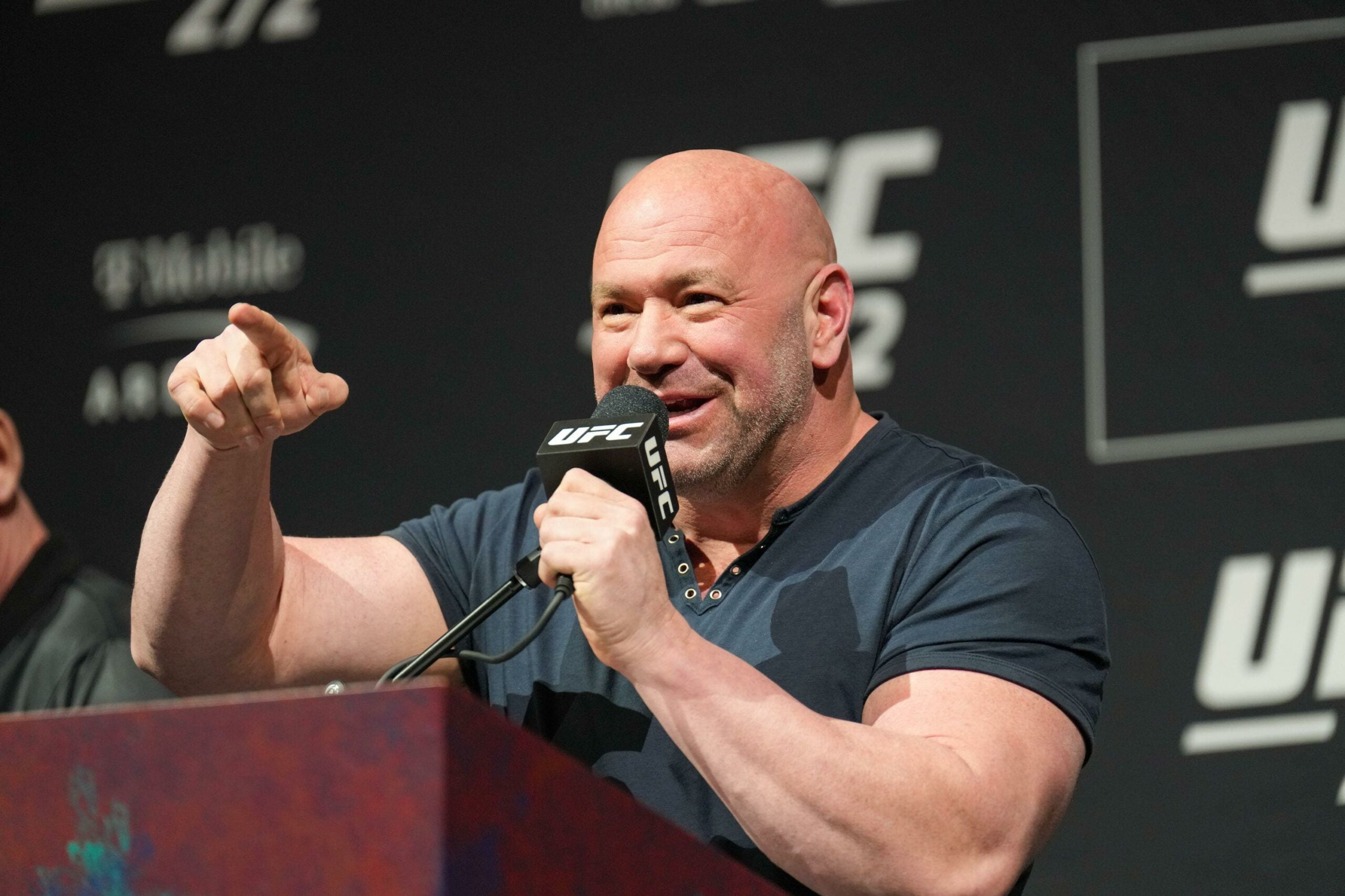 O presidente do UFC, Dana White, vestindo uma camiseta preta, fala ao microfone durante uma coletiva de imprensa, gesticulando com a mão direita enquanto sorri, com um fundo de logotipos do UFC e de uma arena.