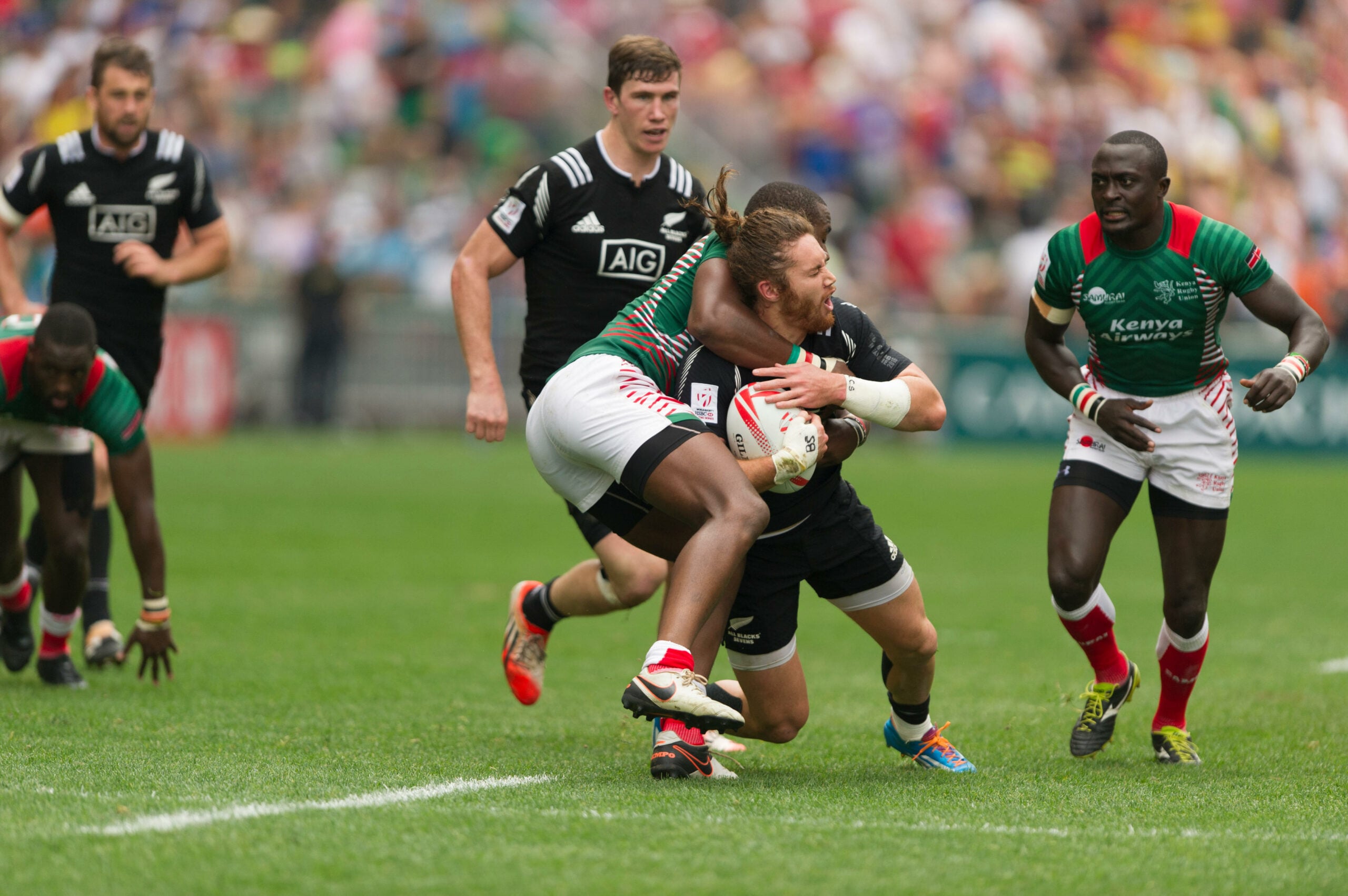 Rugby Sevens: Collins Injera do Quênia, enfrentando Gillies Kaka, da Nova Zelândia. O jogador tenta impedir o outro de avançar com a bola, derrubando-o no chão. Outros dois jogadores estão em campo, ao fundo.