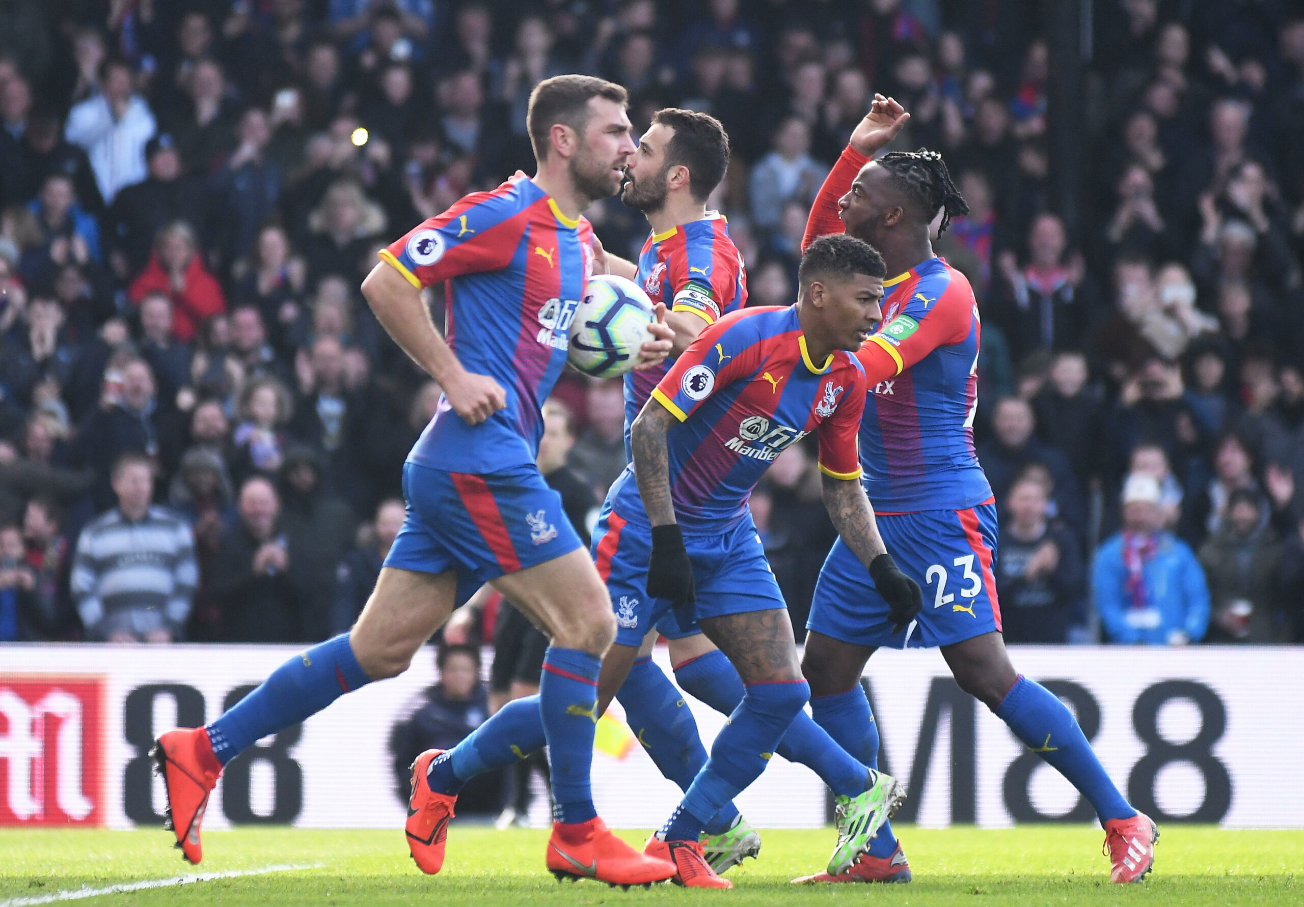 Jogadores do Crystal Palace comemoram um gol com entusiasmo, vestindo seus uniformes vermelhos e azuis vibrantes, cercados por uma multidão animada de torcedores no estádio.